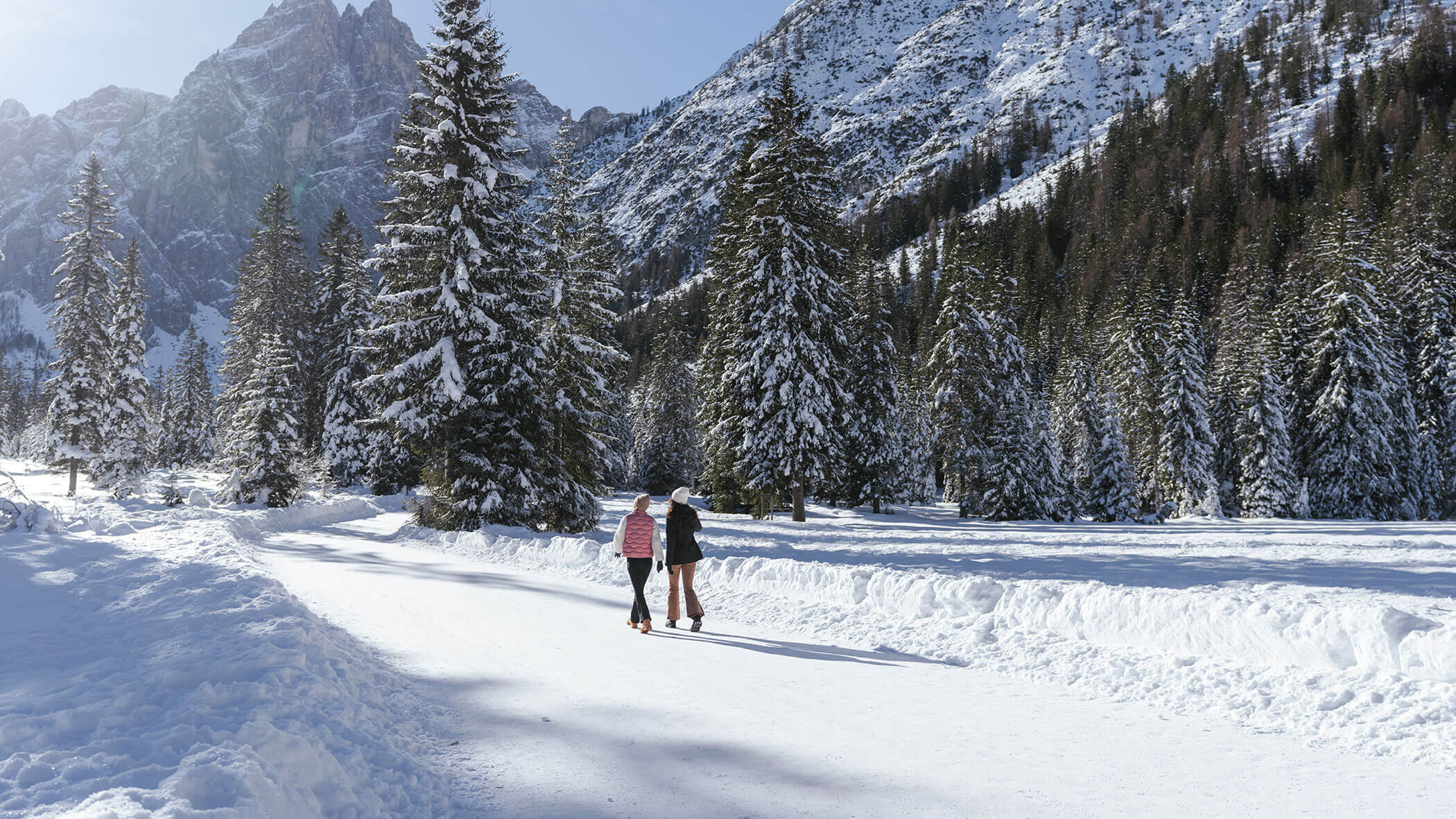 Two people ice skating on a snowy path surrounded by pine trees and snow-covered mountains.