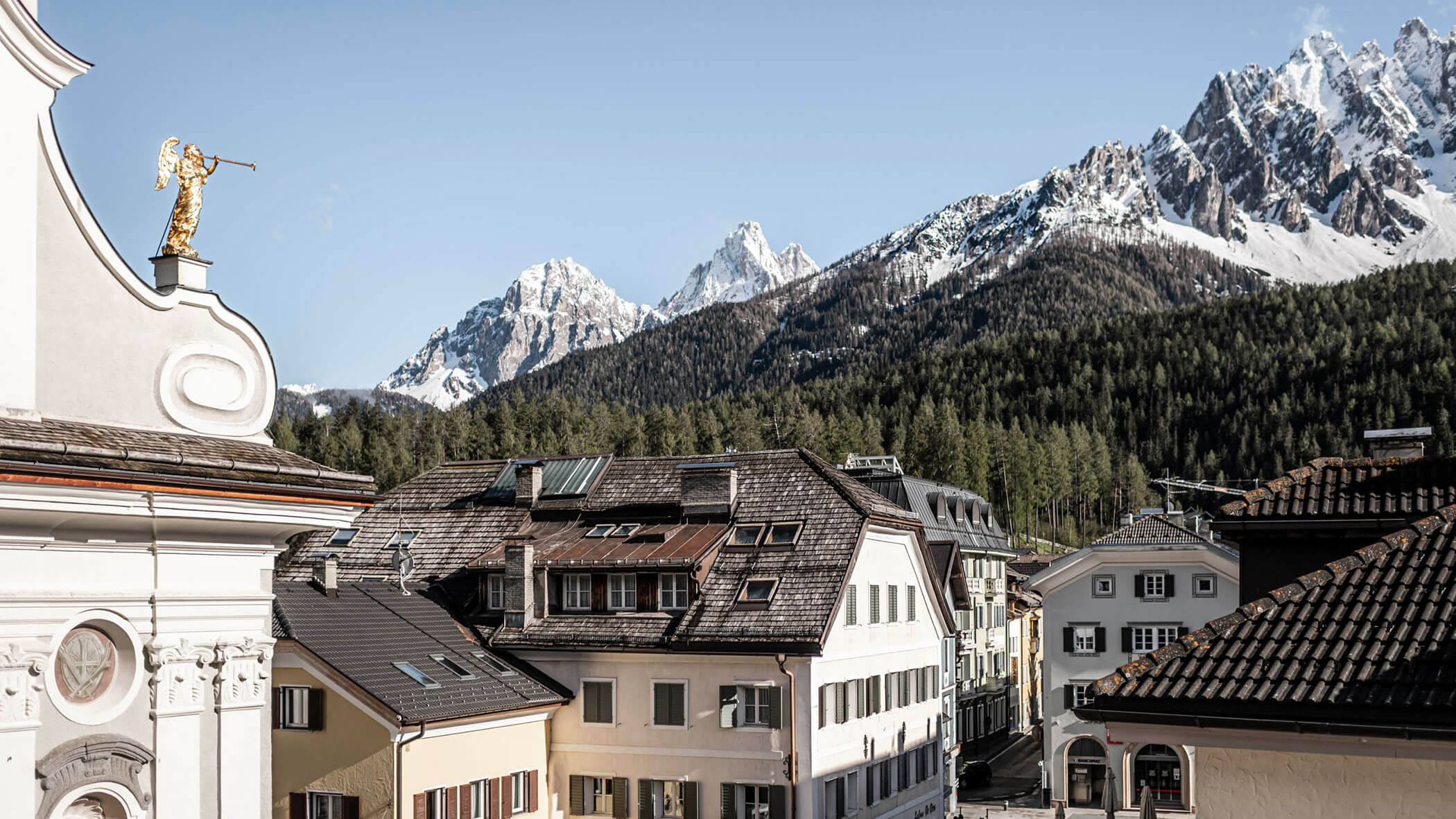 Rooftops of alpine village with snowy mountains and pine forest in the background under a clear sky.