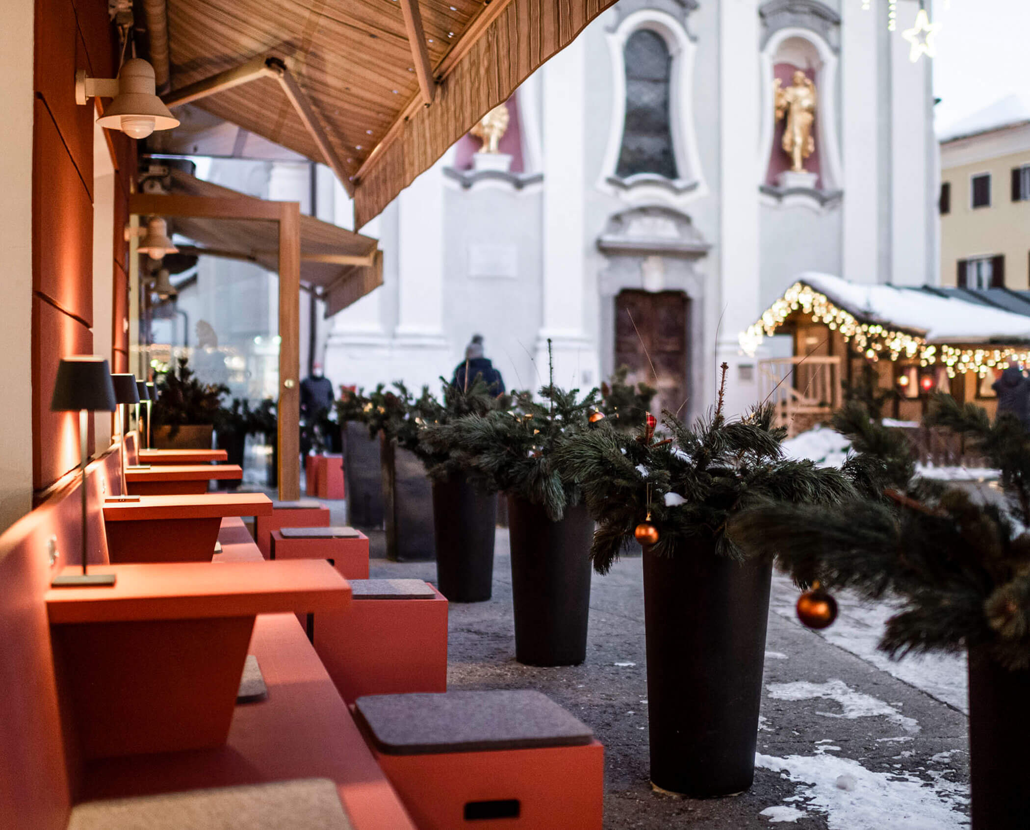 Outdoor seating area with red benches and pine decorations, with a church and festive lights in the background.
