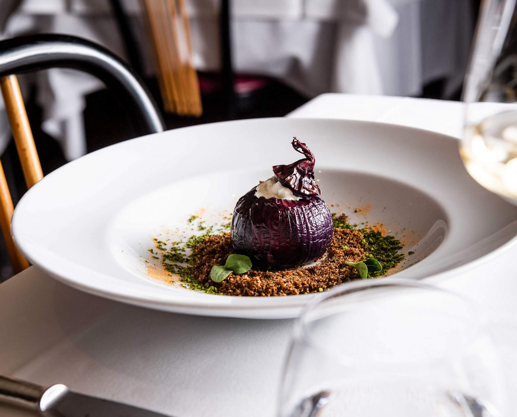 A stuffed roasted red onion is plated on a bed of crumbs and herbs in a white bowl on a restaurant table.