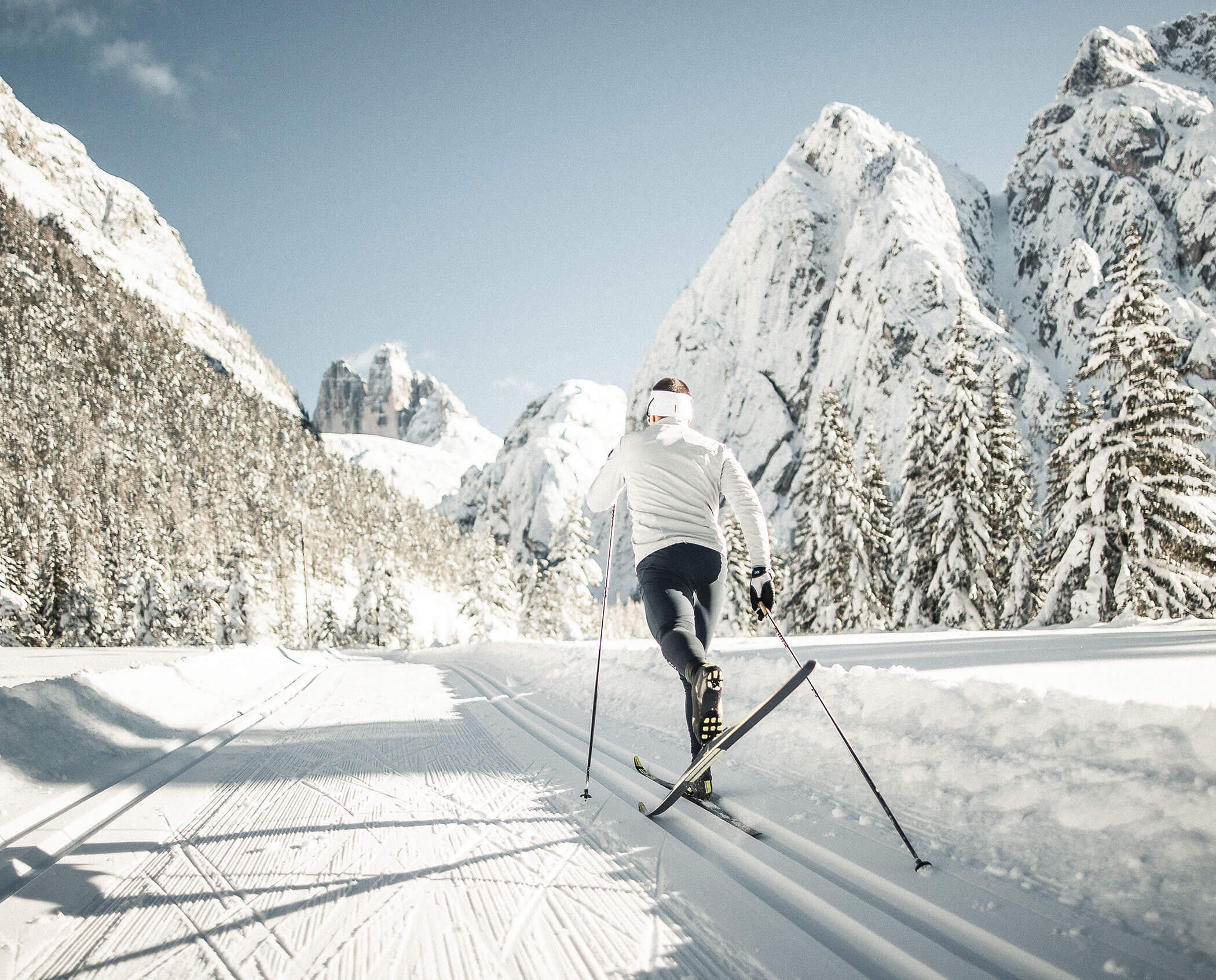 A person cross-country skiing on a snowy trail with mountains and pine trees in the background.