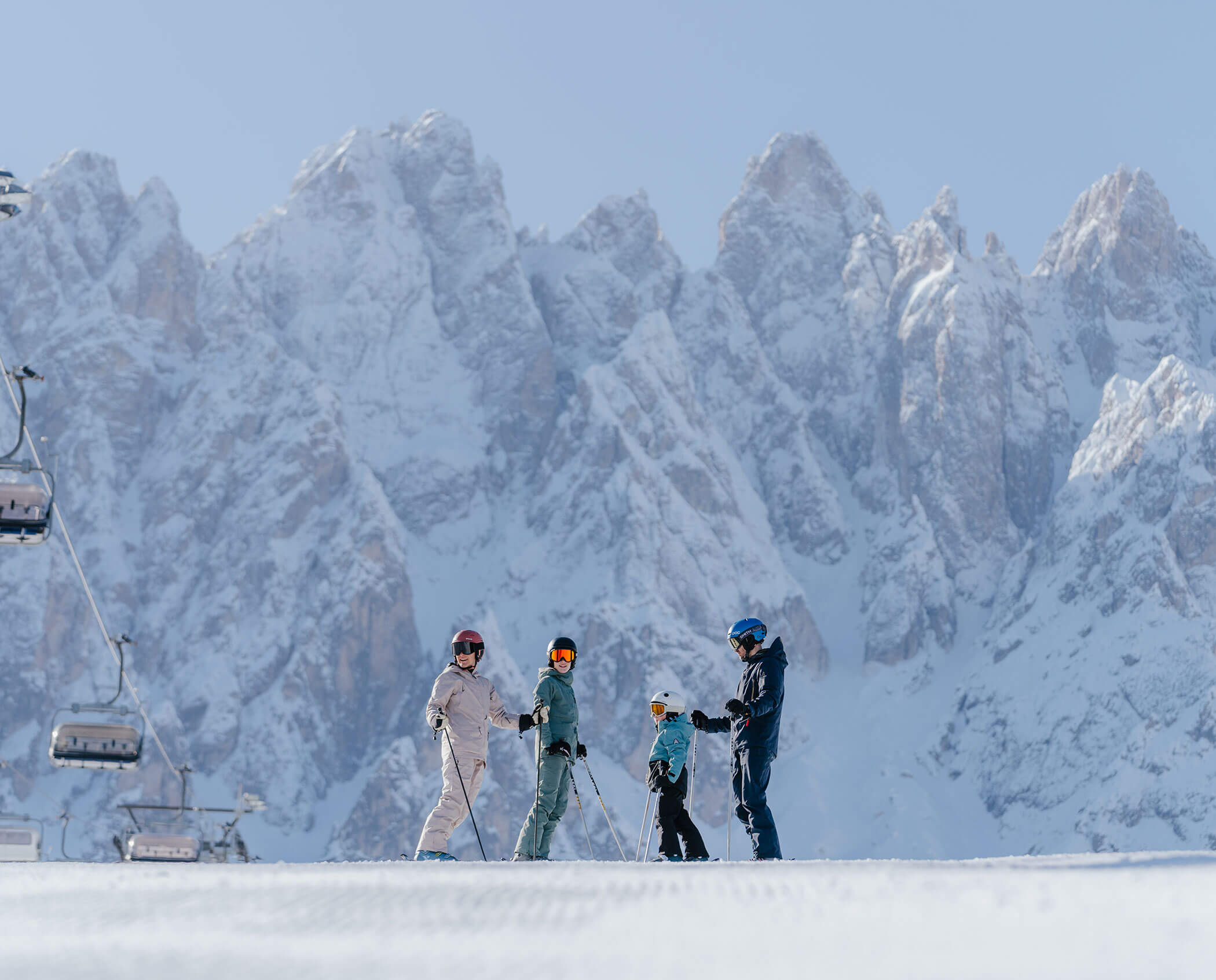 Four skiers standing on a snowy slope with tall, rocky mountains and ski lifts in the background.