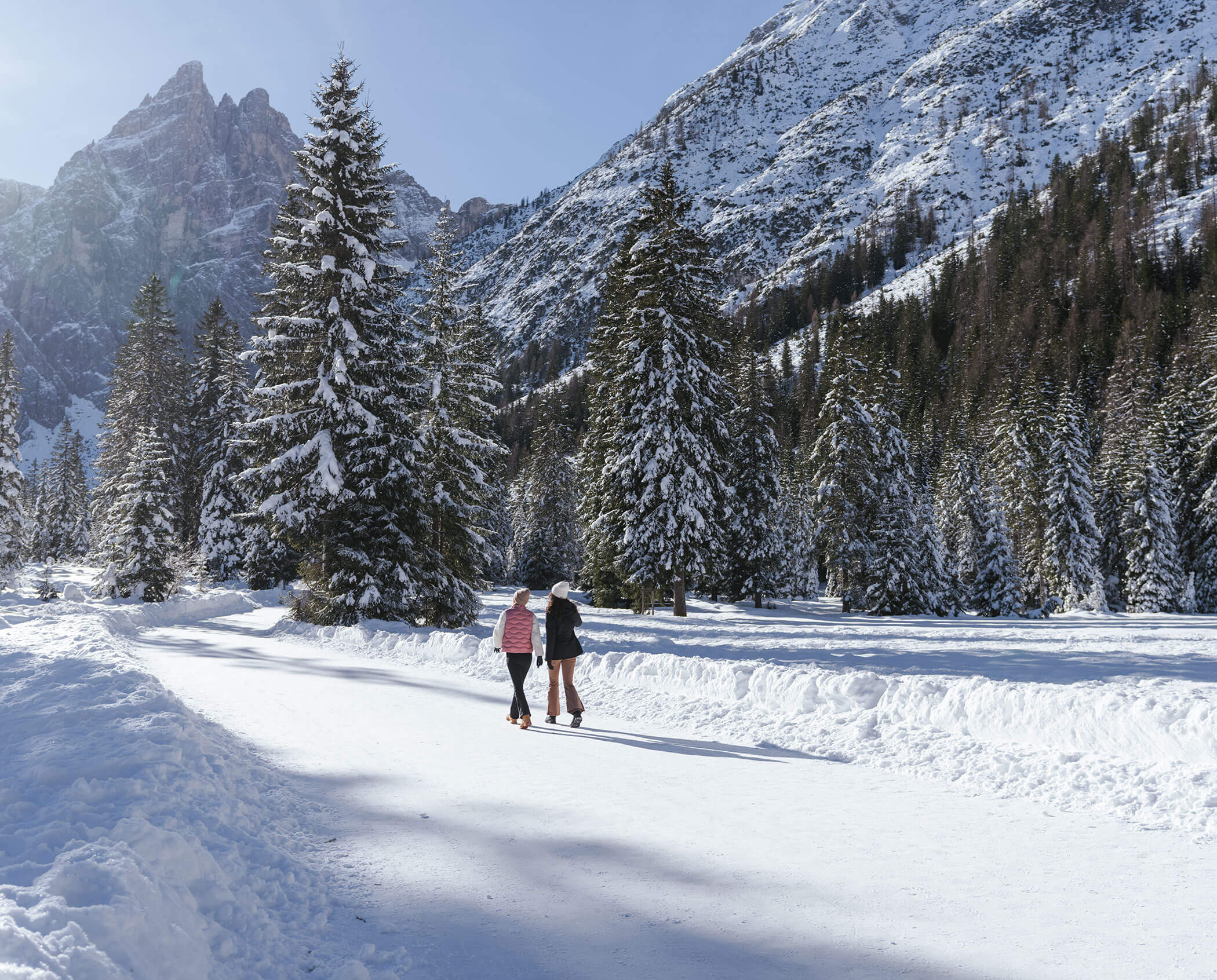 Two people ice skating on a snowy path surrounded by pine trees and mountains on a sunny winter day.