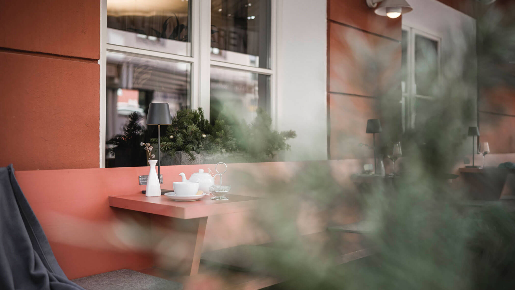 A cozy outdoor café table with tea set, bench seating, and plants, partially obscured by greenery.