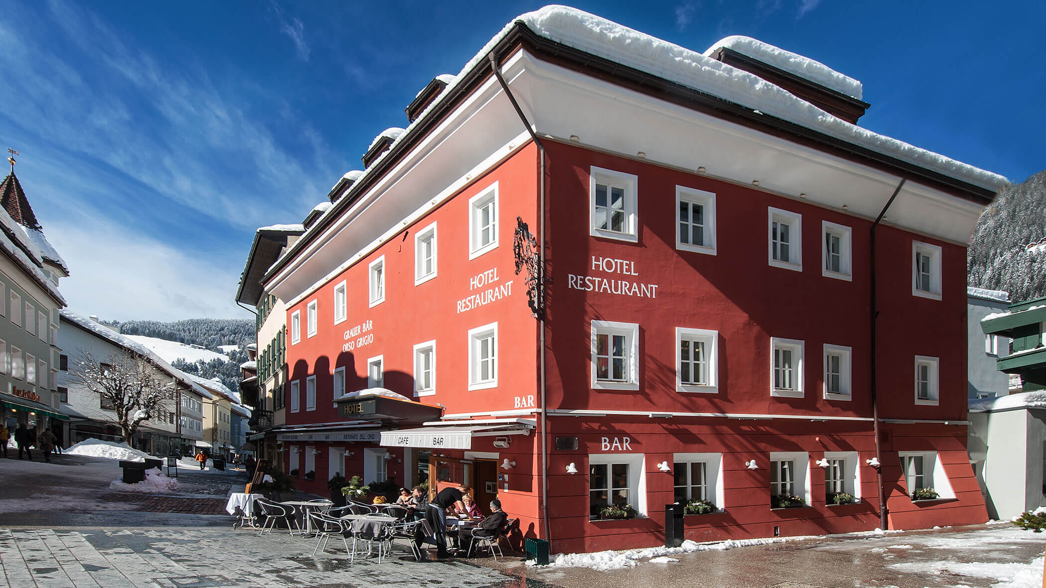 A red hotel and restaurant with outdoor seating on a snowy street under a clear blue sky.