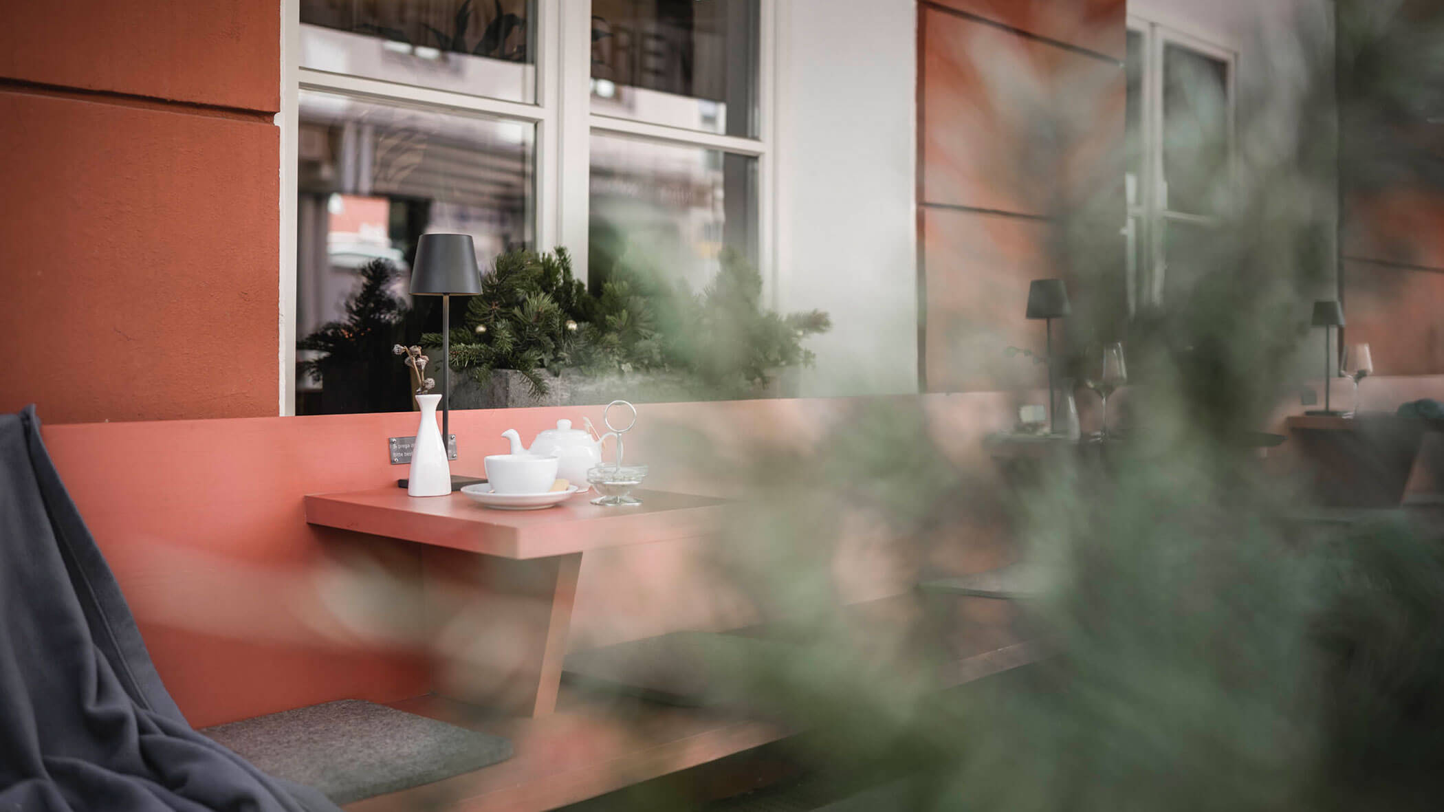 Outdoor café table with tea set and lamp, partially obscured by green foliage in the foreground.