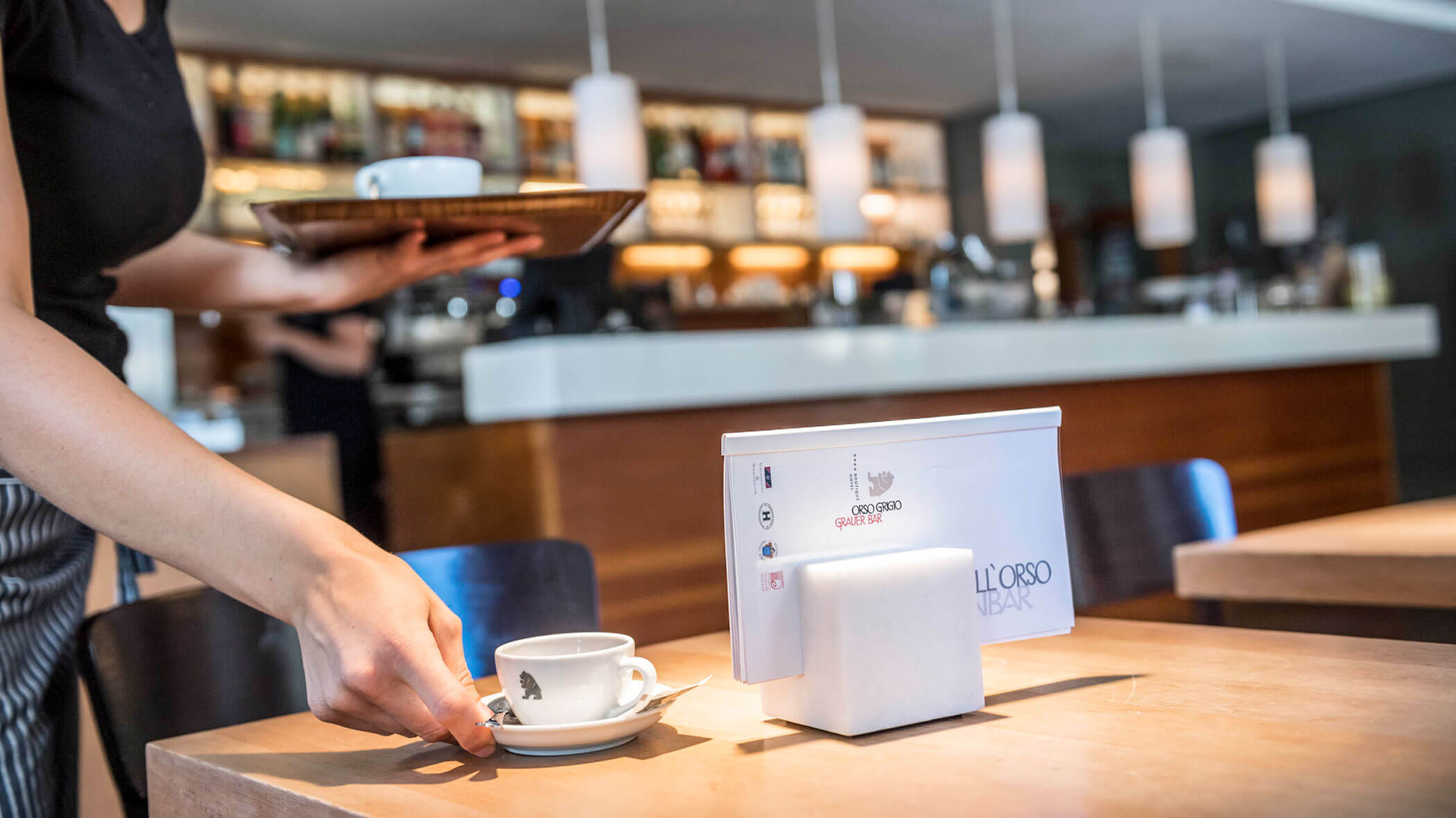 A server sets a cup of coffee on a wooden table in a modern café with a bar in the background.
