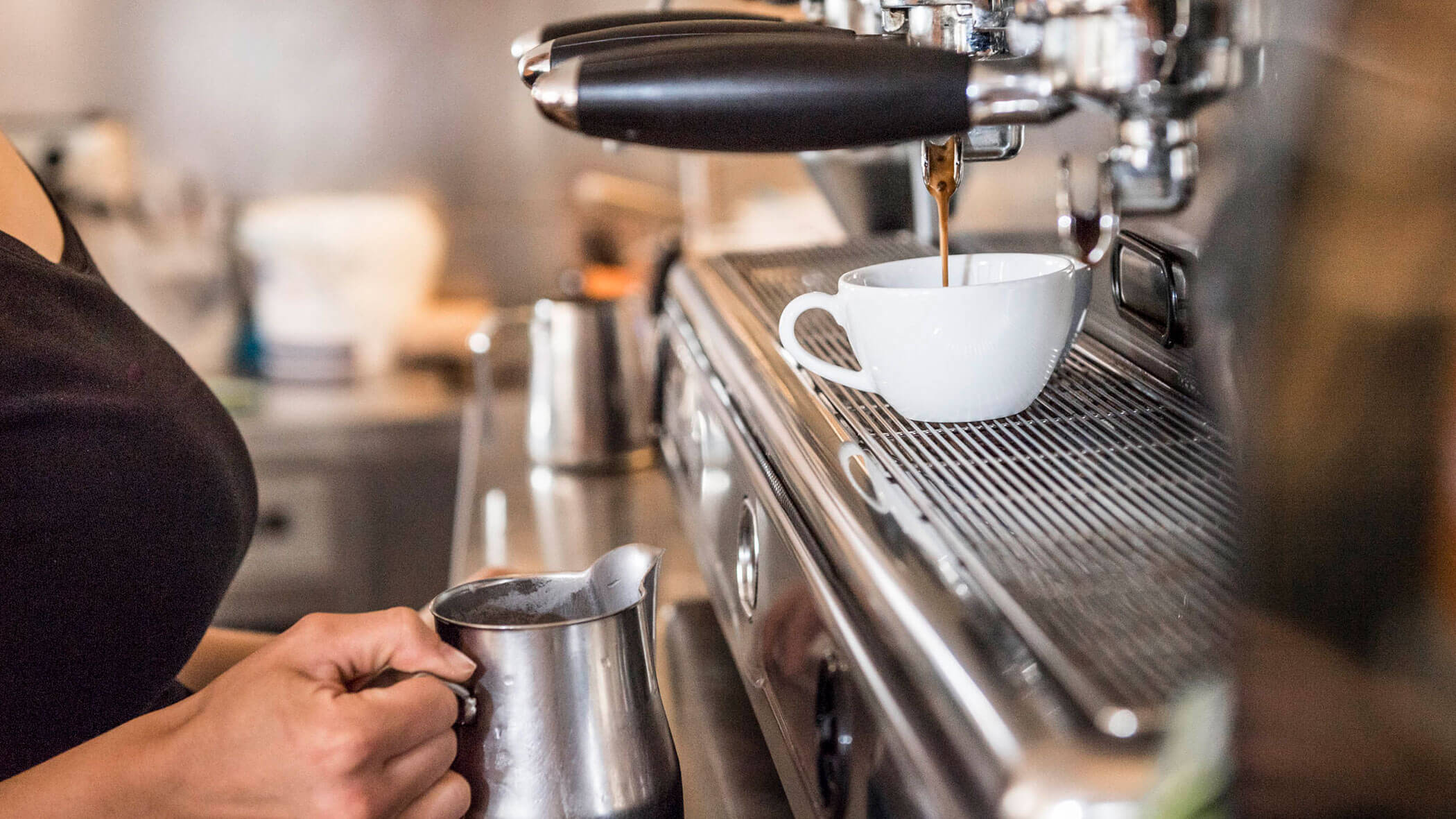 A person making coffee with an espresso machine, holding a milk frothing pitcher next to a white cup.