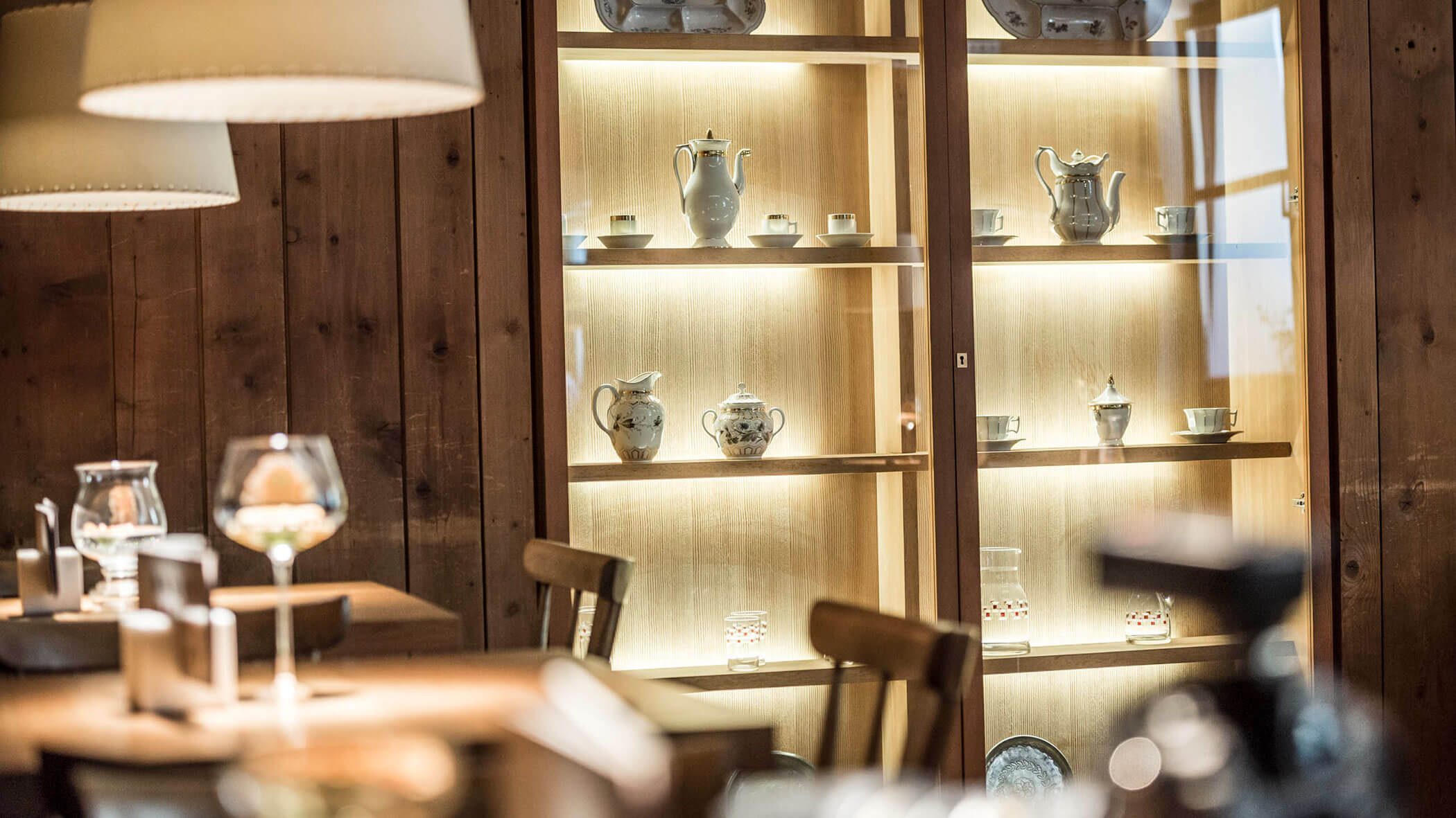 A glass cabinet displays teapots and cups in a cozy, warmly lit wooden dining room.