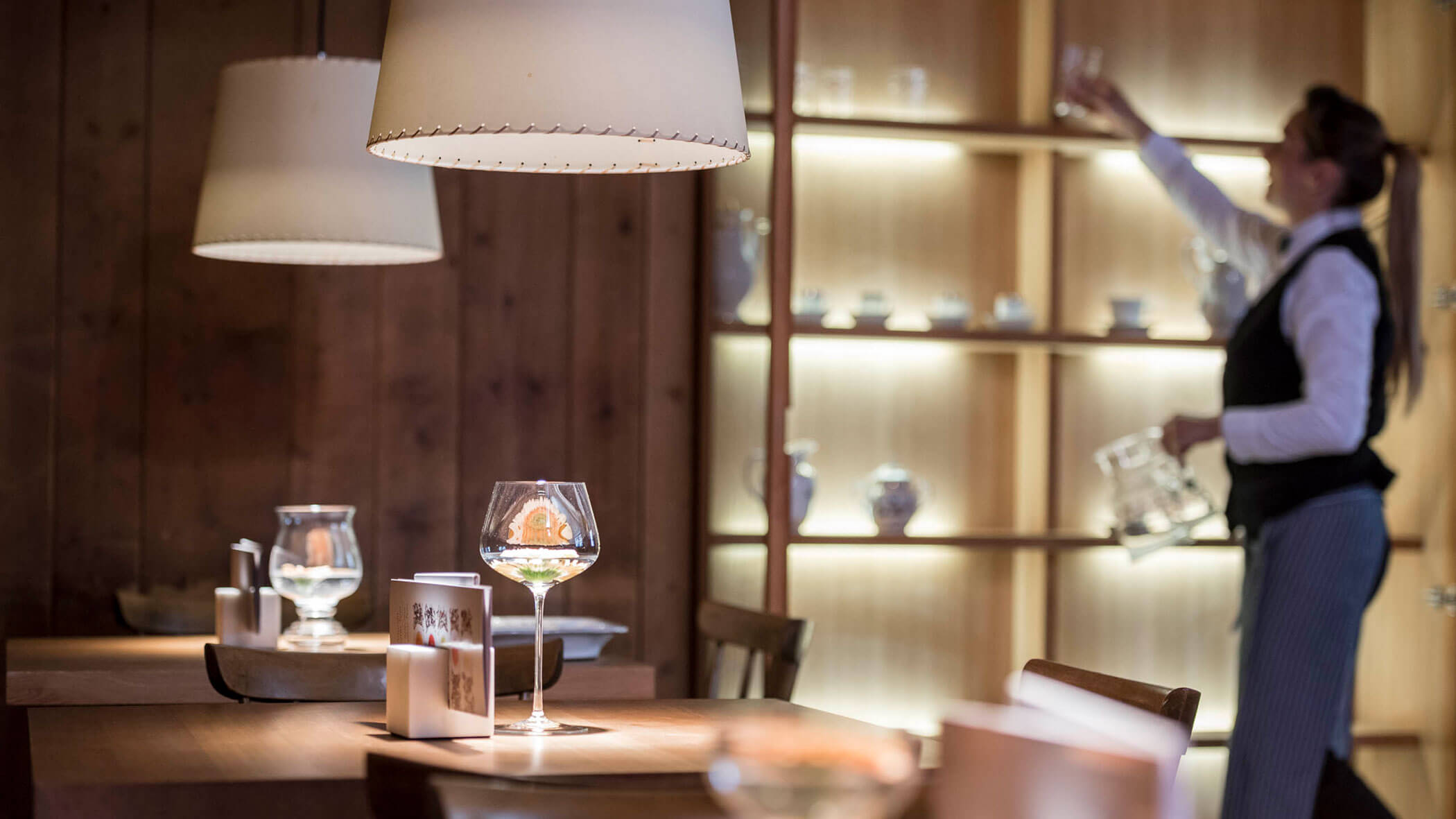 A server arranges glassware on a shelf in a cozy, softly lit restaurant with wooden decor.