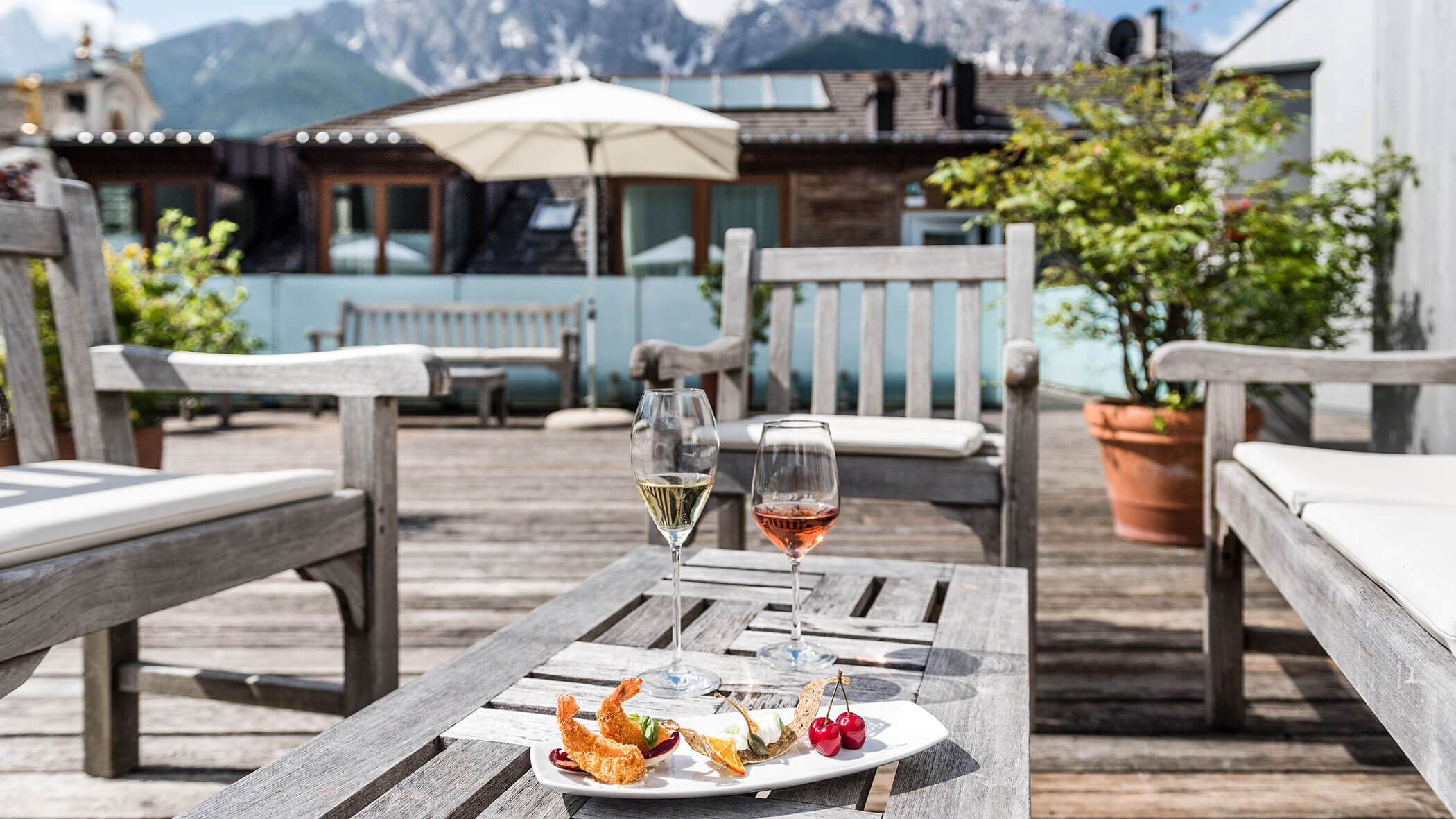 Due bicchieri di vino e un piatto da dessert su un tavolo di legno del patio, con montagne ed edifici sullo sfondo.