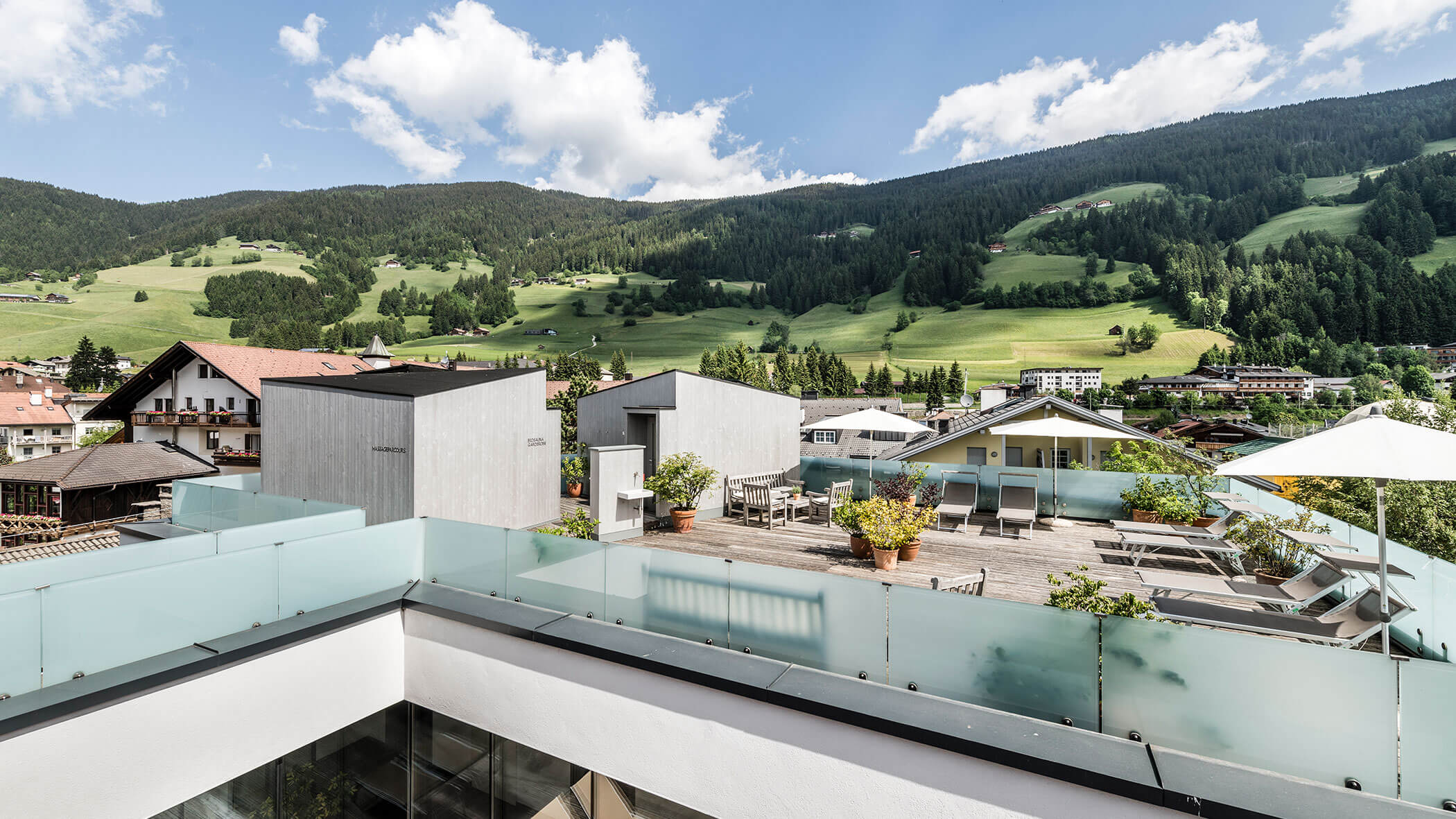 Rooftop terrace with lounge chairs and potted plants, overlooking green hills and a mountain village.