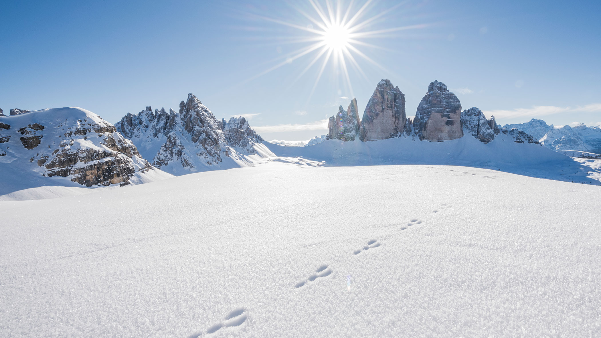 Snowy mountain peaks with sunburst overhead and footprints crossing a pristine snowfield in the foreground.