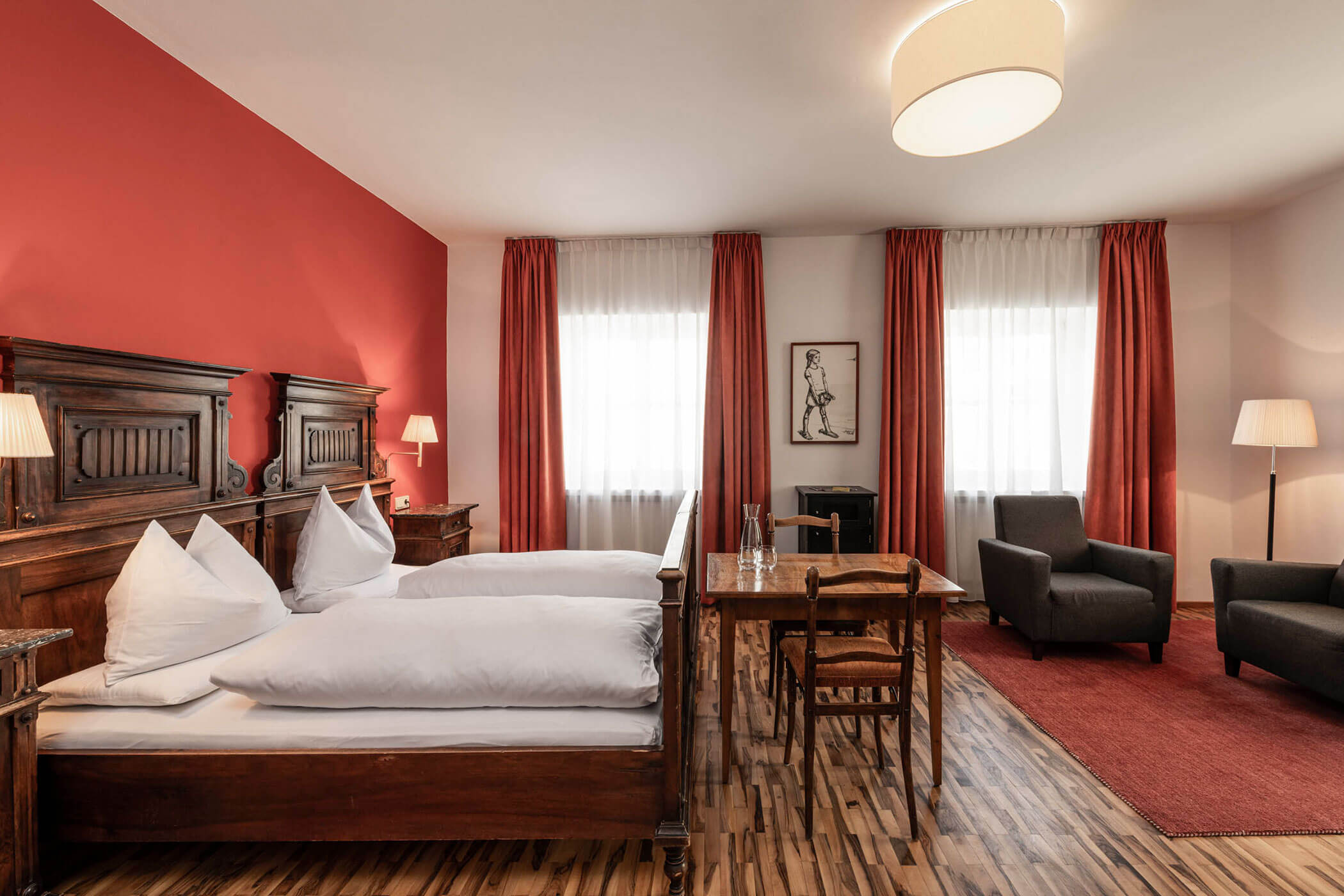A cozy hotel room with twin beds, wooden furniture, red curtains, and a small sitting area.
