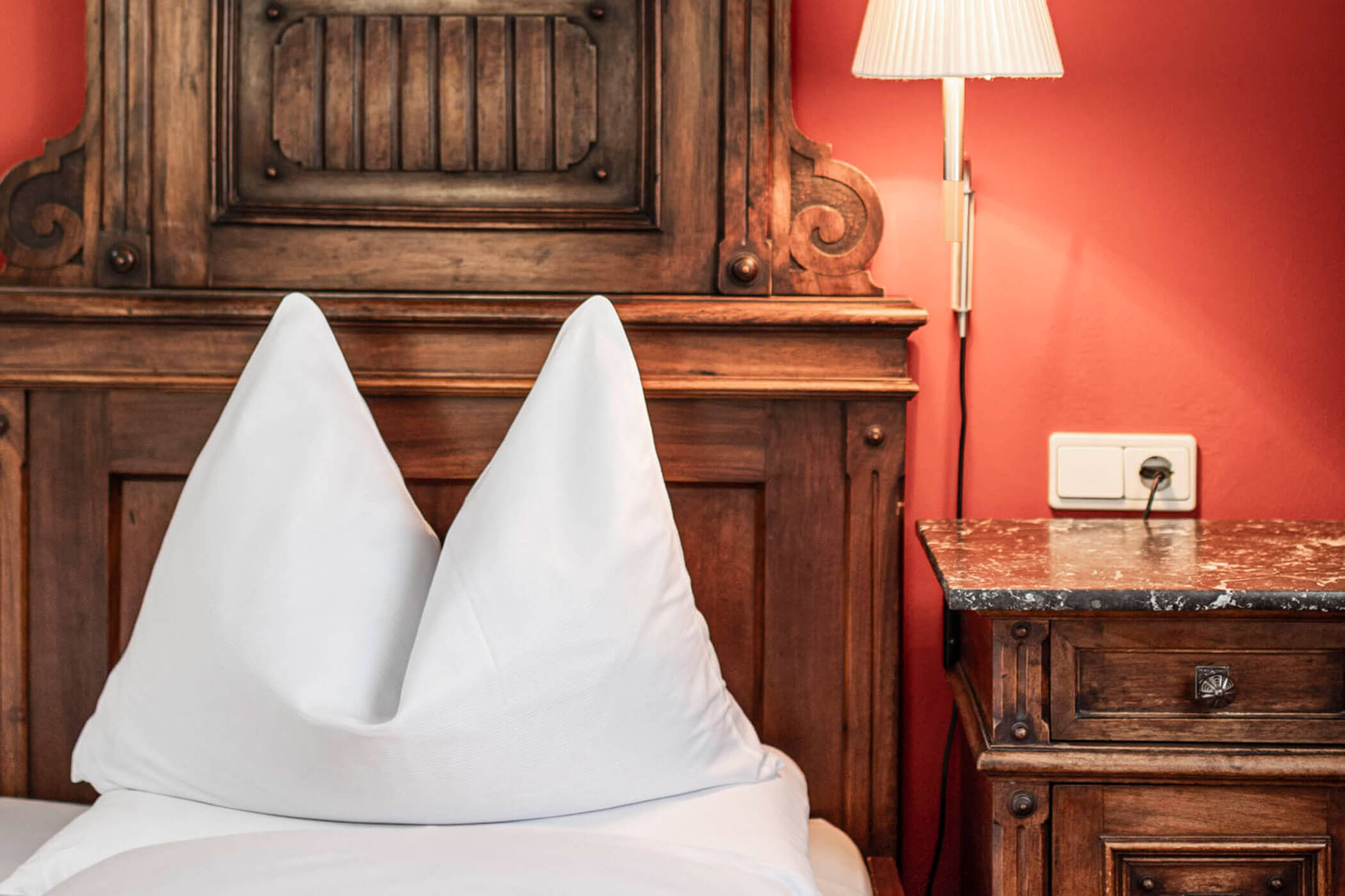 White pillows on a bed with an ornate wooden headboard and a wooden nightstand against a red wall.