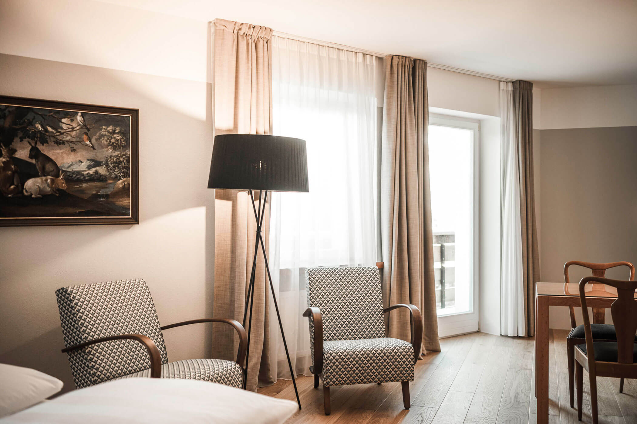 Bright room with two patterned armchairs, a floor lamp, large window, and a framed painting on the wall.