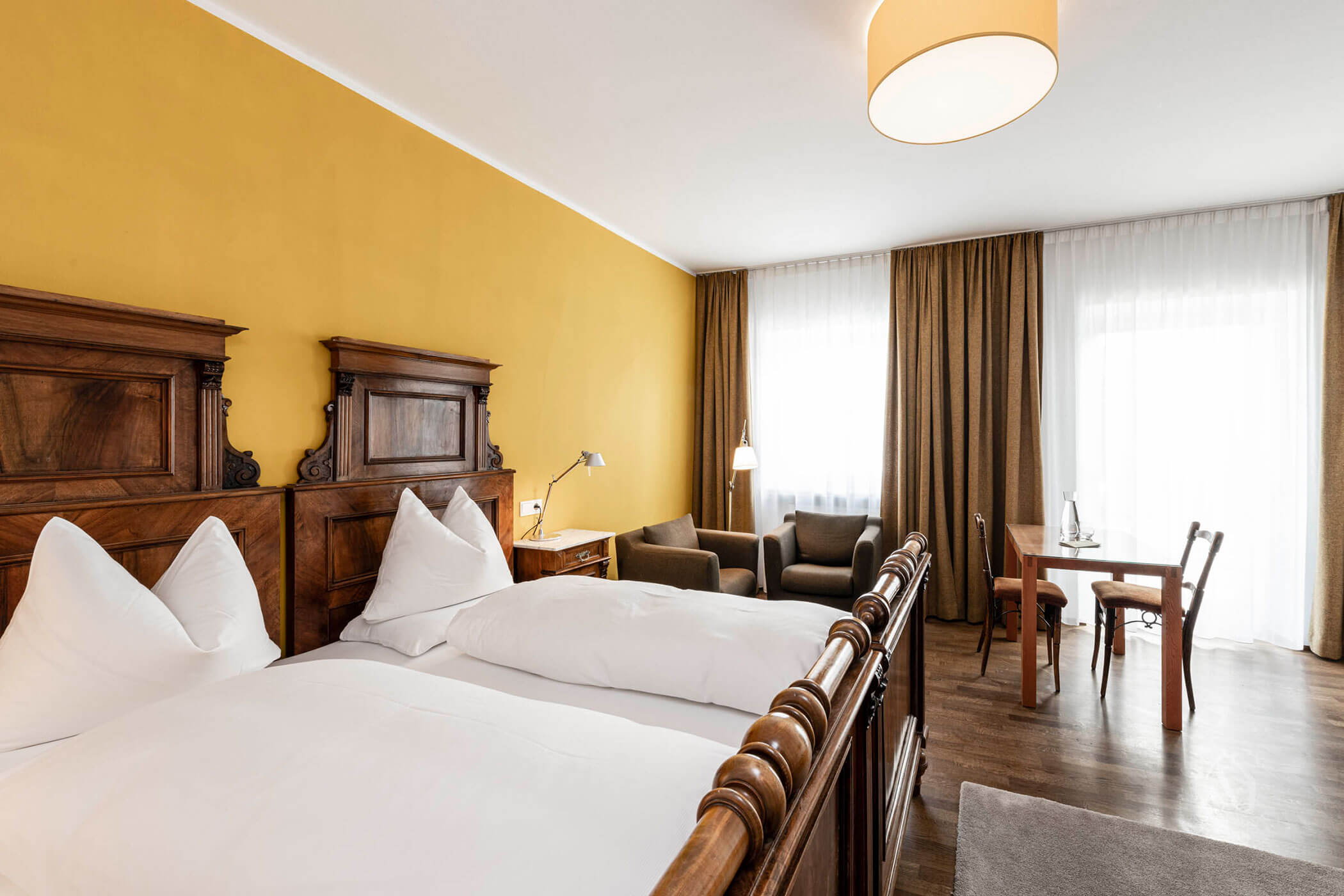 Hotel room with two wooden beds, yellow wall, seating area, table, chairs, and large windows with curtains.