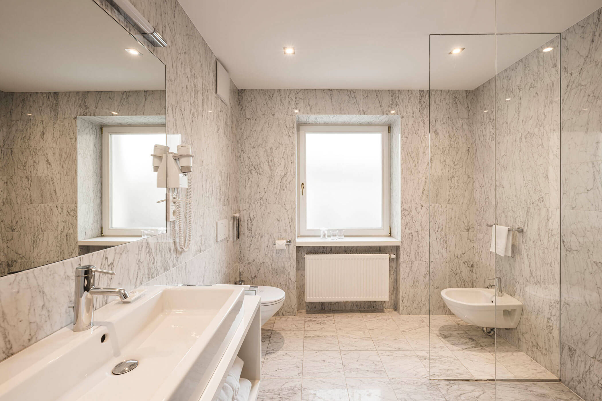 Modern marble bathroom with large mirror, white sink, toilet, bidet, and a window letting in natural light.