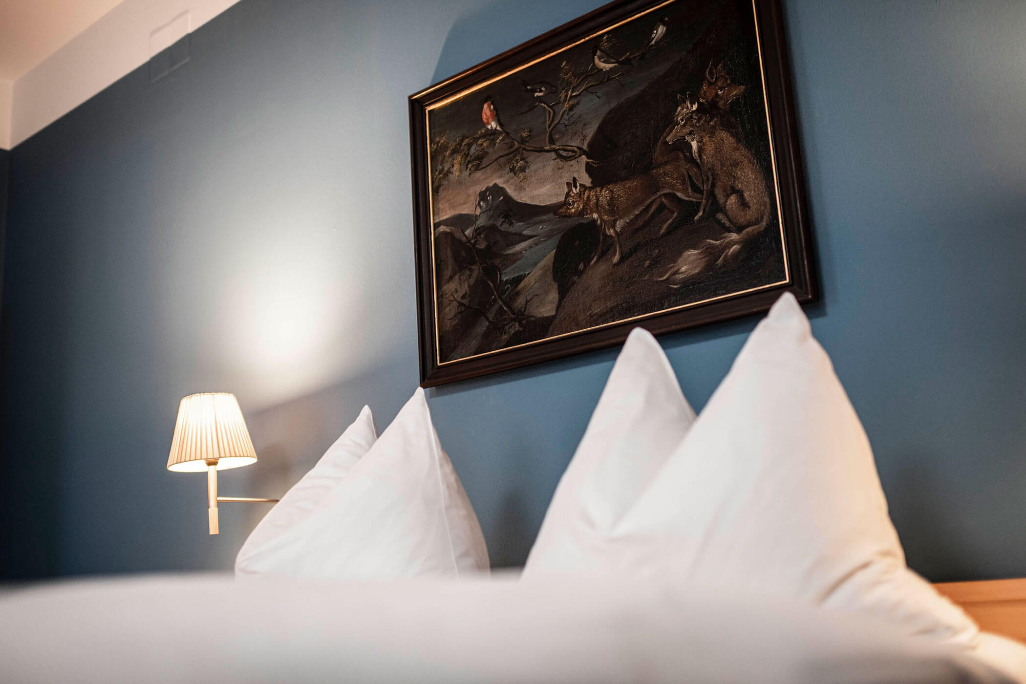 Two white pillows on a bed, a wall lamp, and a framed animal painting on a blue wall.