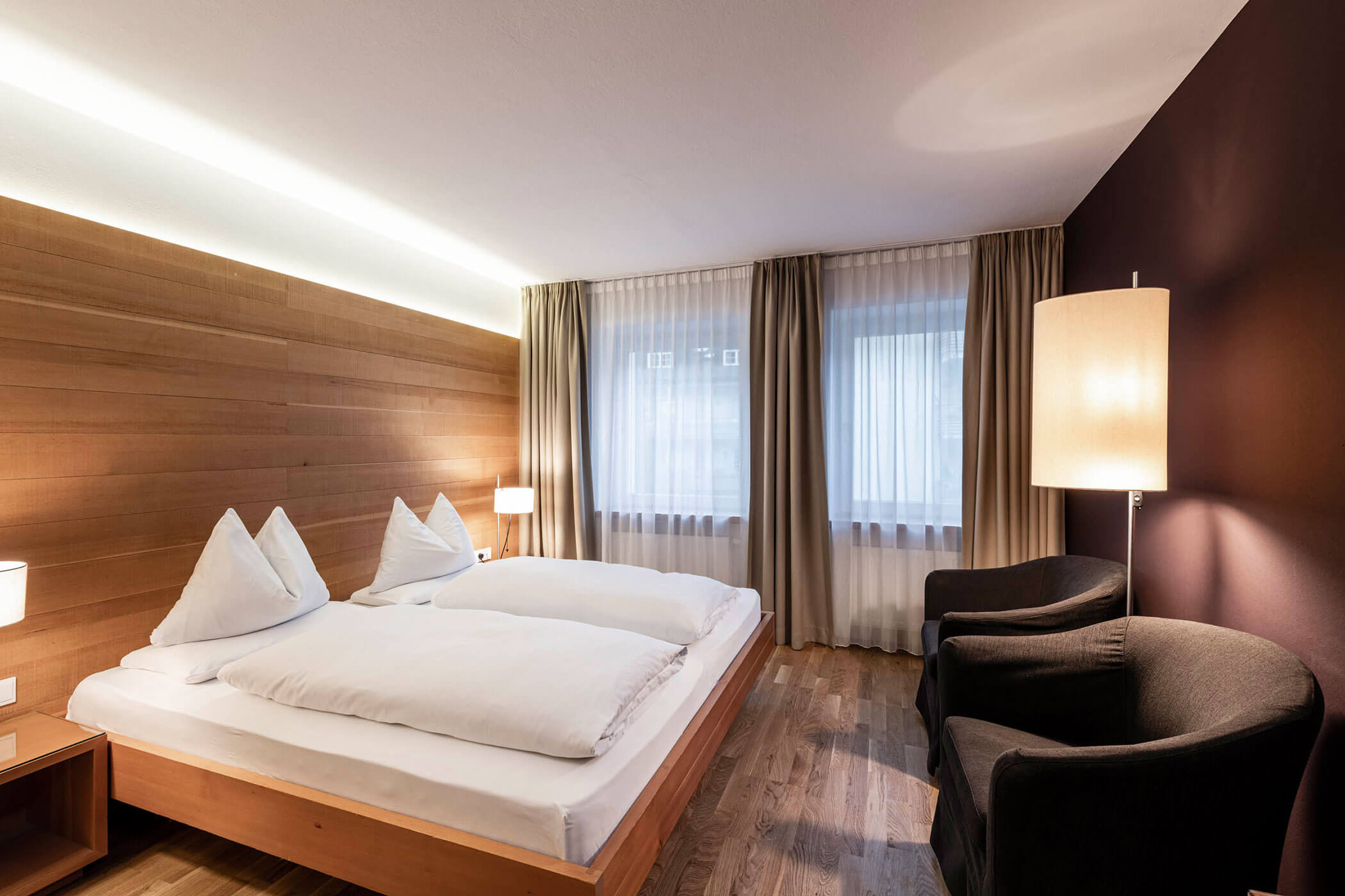 Modern hotel room with a large bed, two chairs, wooden floors, and warm lighting from lamps.