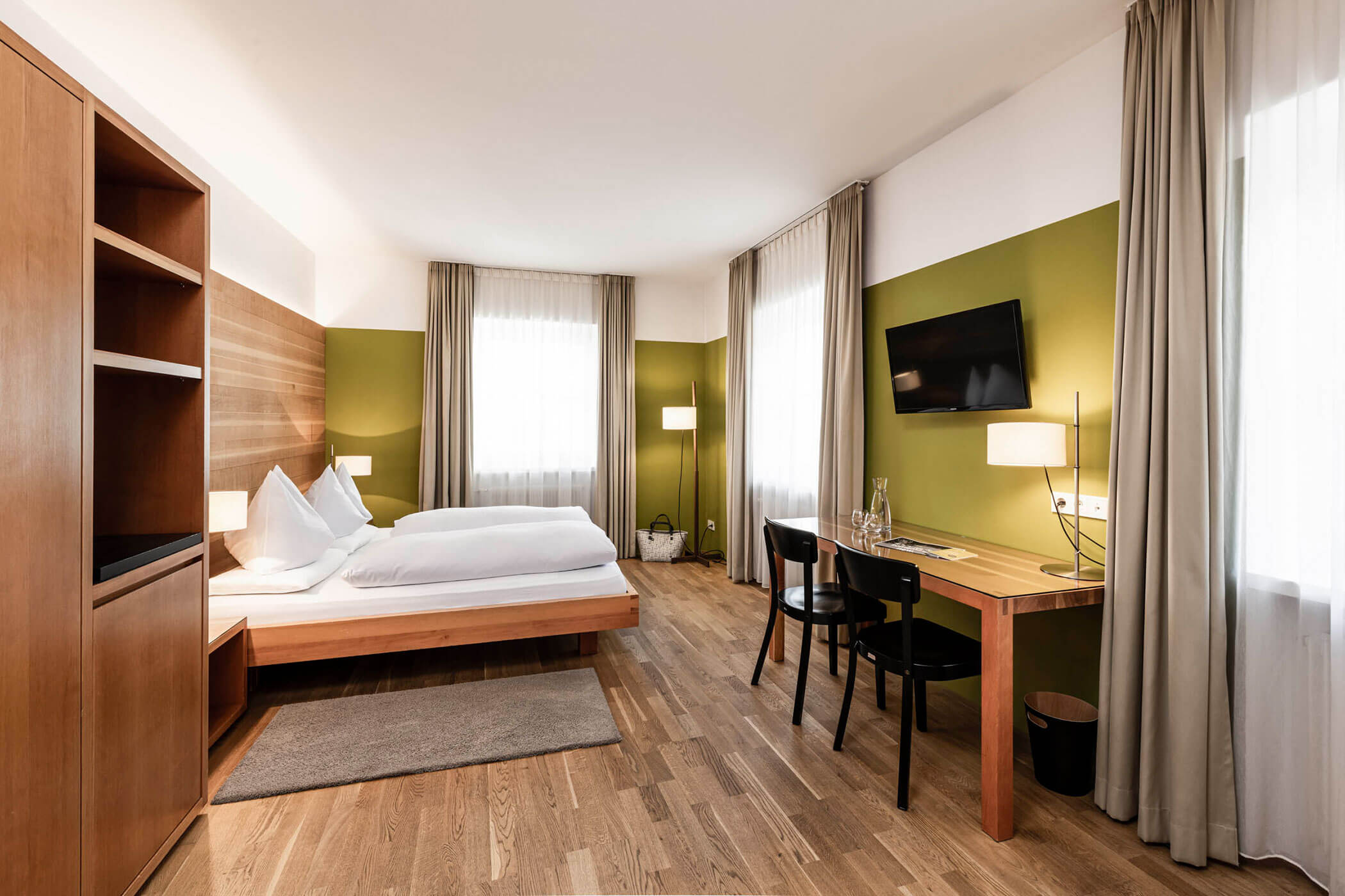 Modern hotel room with a double bed, desk, TV, large windows, and wooden flooring.