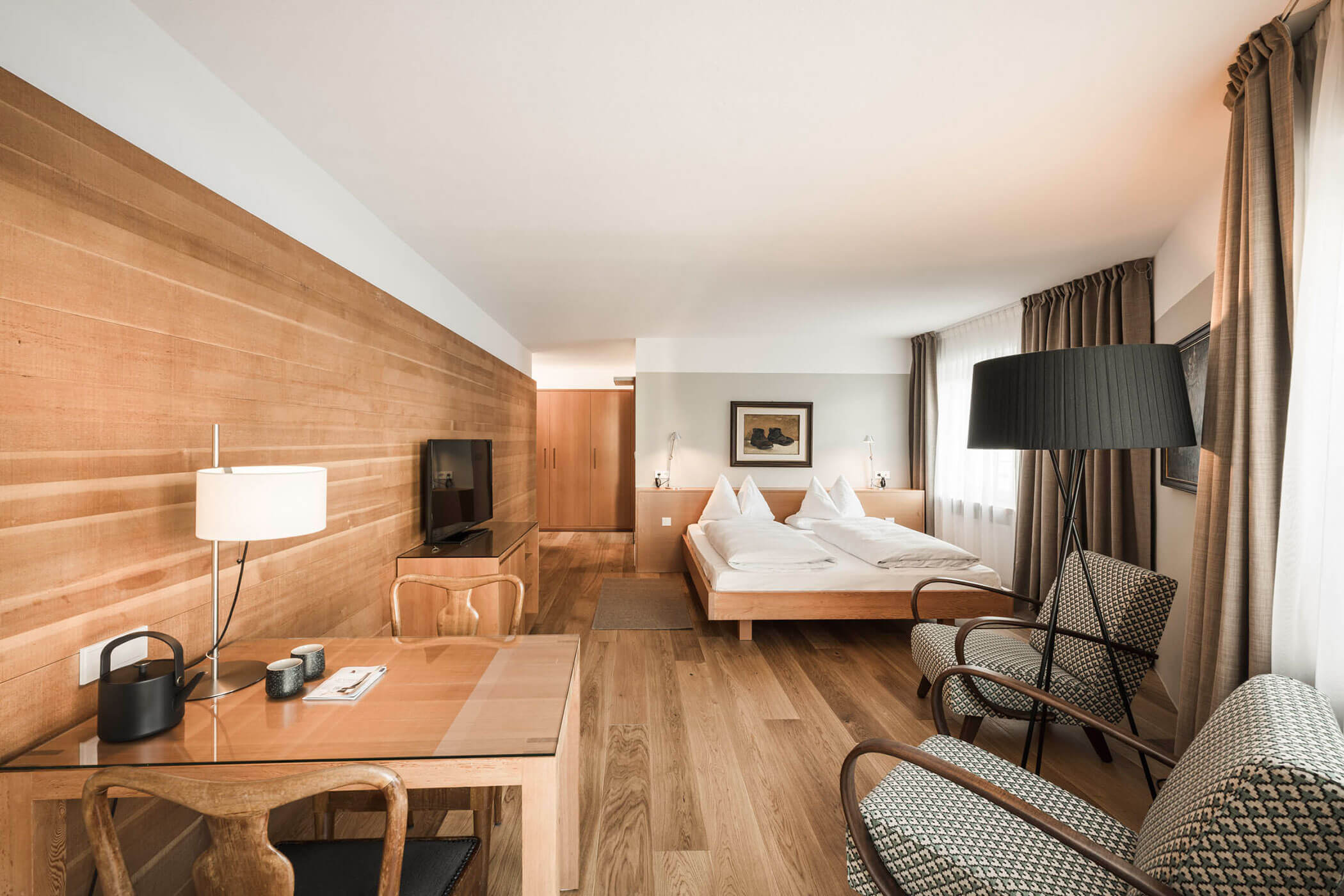 Modern hotel room with wood floors, a double bed, desk, armchairs, TV, and large windows with curtains.