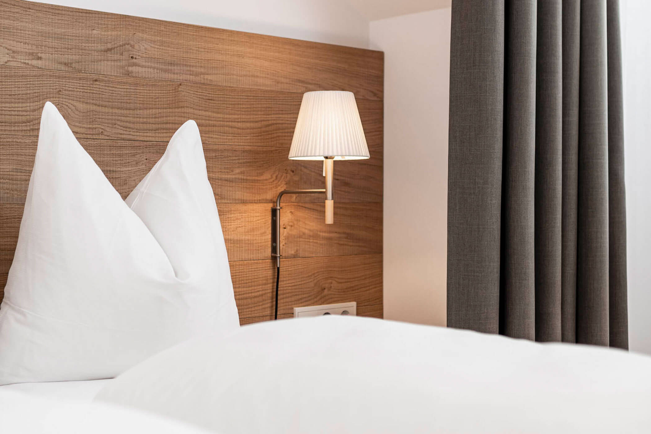 White pillows and bedding on a neatly made bed with a wood headboard and wall-mounted lamp.