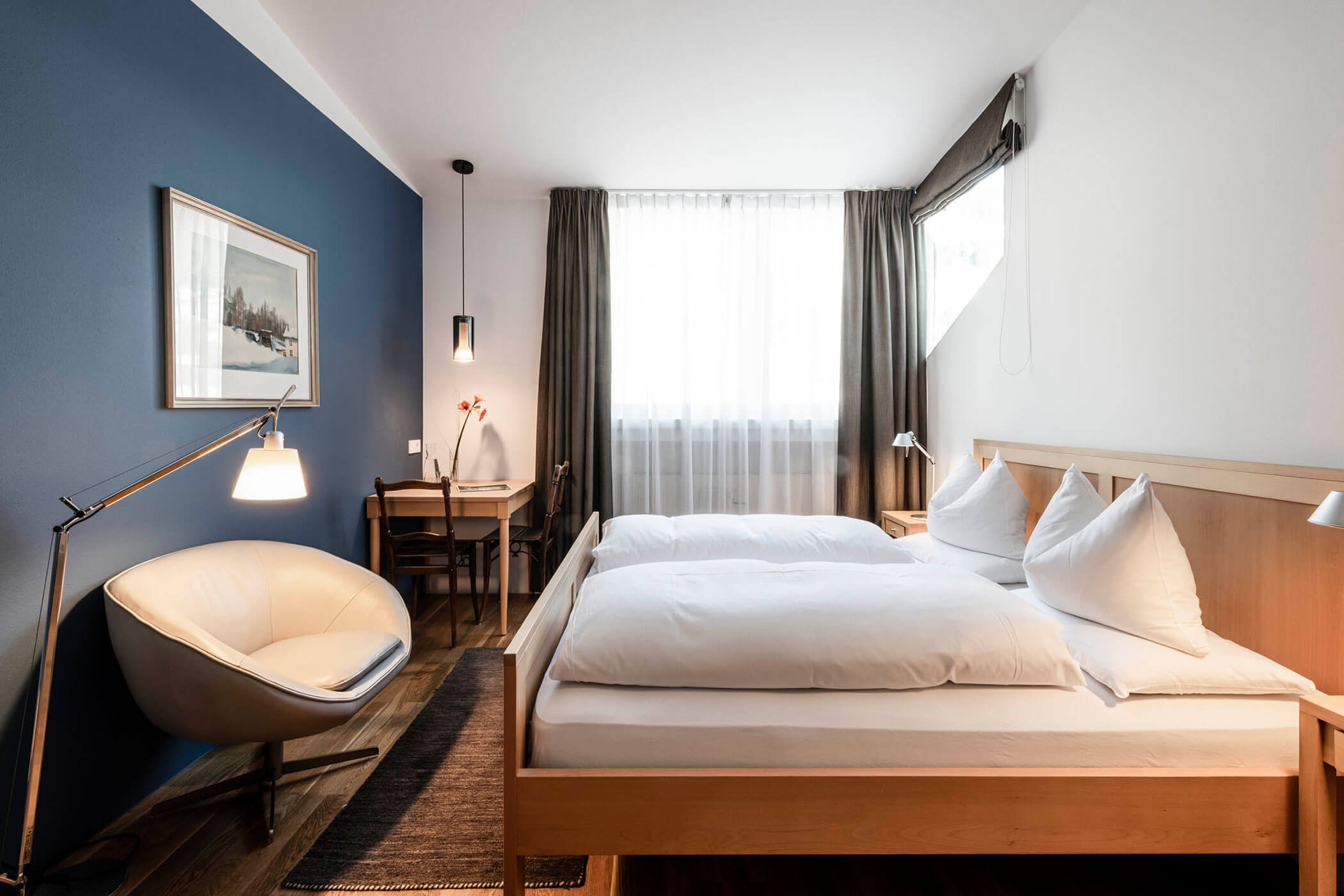 Modern hotel room with a double bed, armchair, desk, blue accent wall, and large window with curtains.