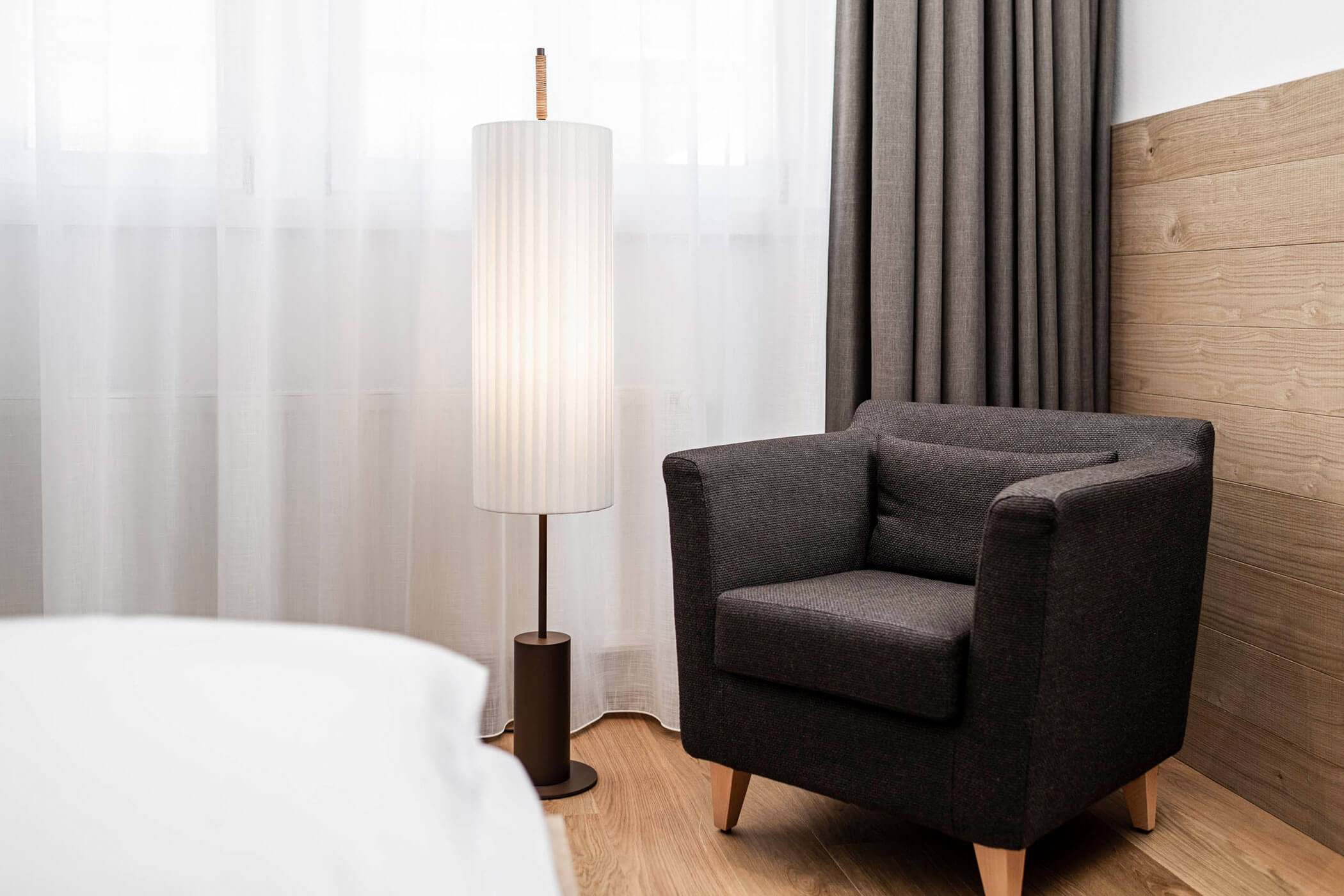 Dark armchair and tall floor lamp beside a window with sheer curtains and gray drapes in a cozy room.
