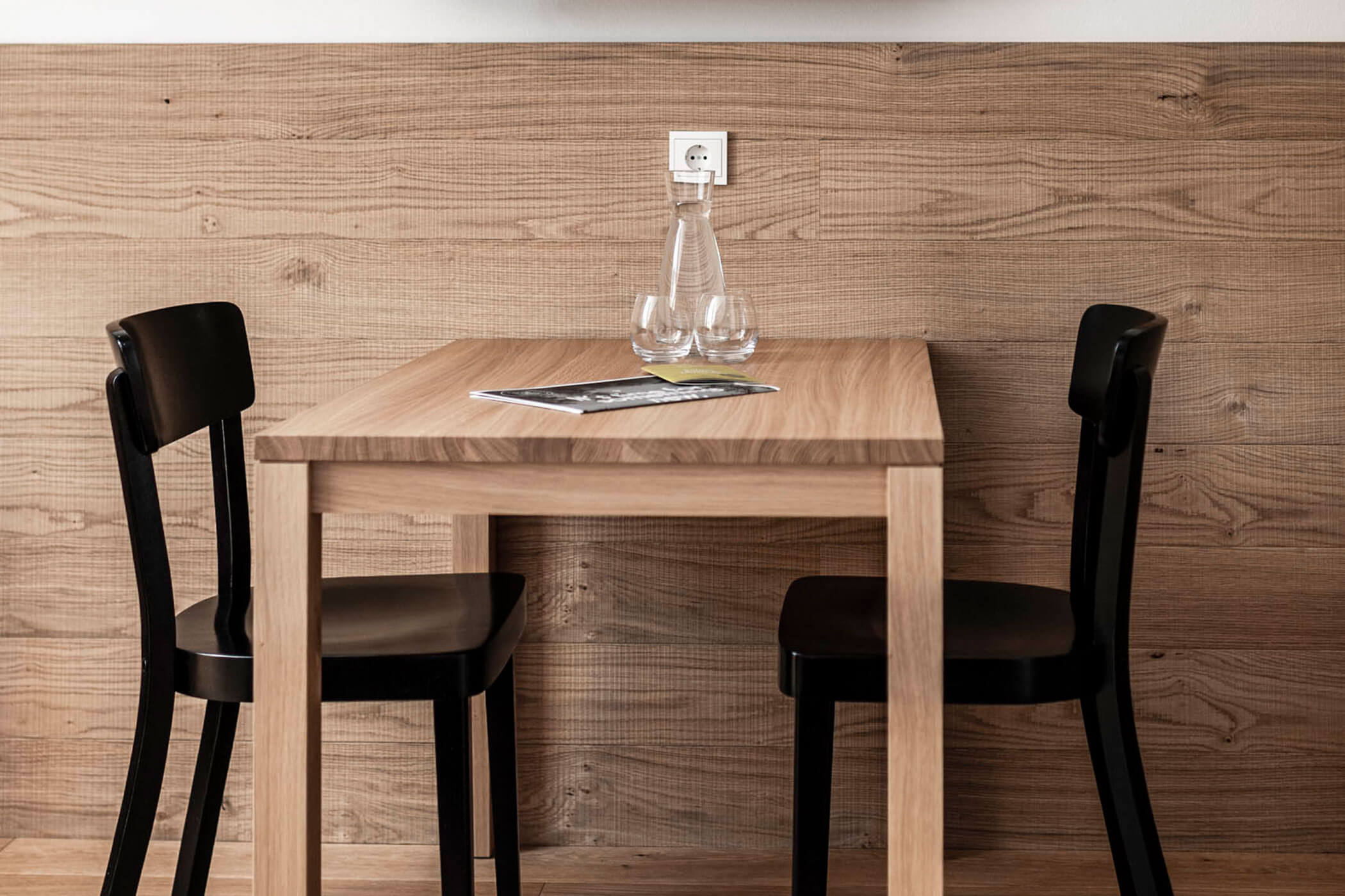 A small wooden table with two black chairs, a glass bottle, two glasses, and a magazine on top.