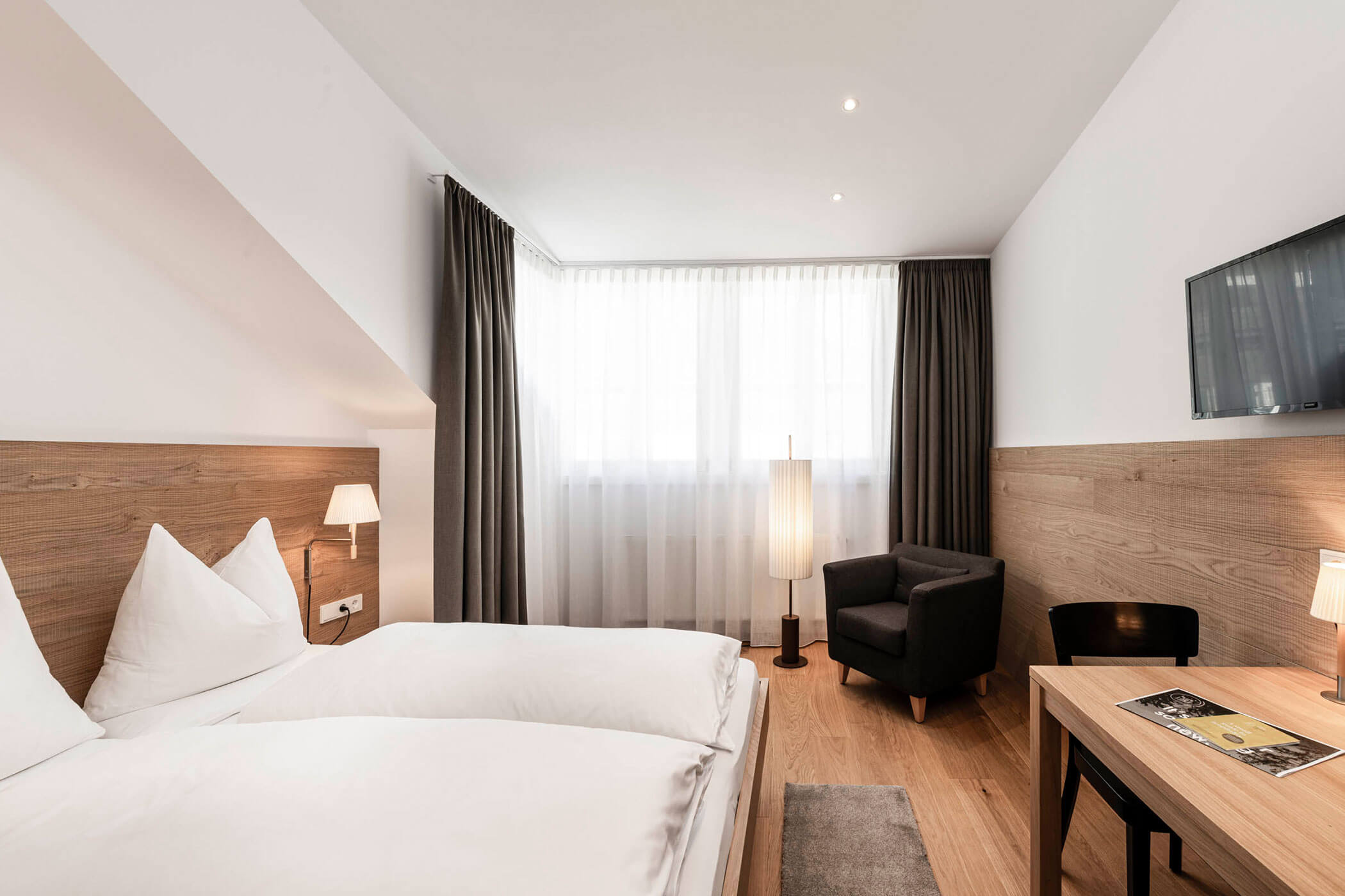Bright hotel room with twin beds, armchair, desk, TV, wood accents, and large window with curtains.