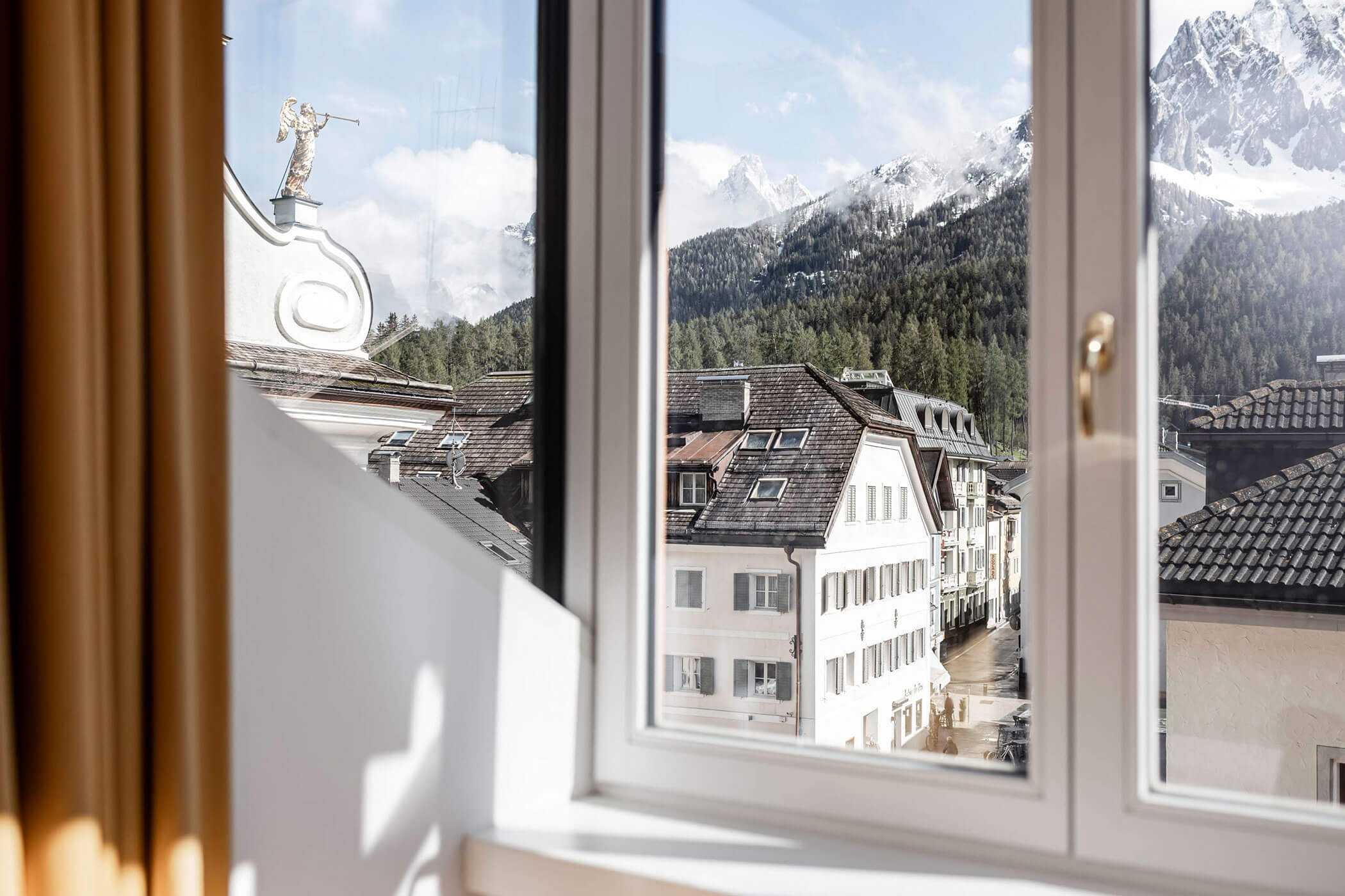 View through a window of alpine buildings, forest, and snow-capped mountains under a clear sky.