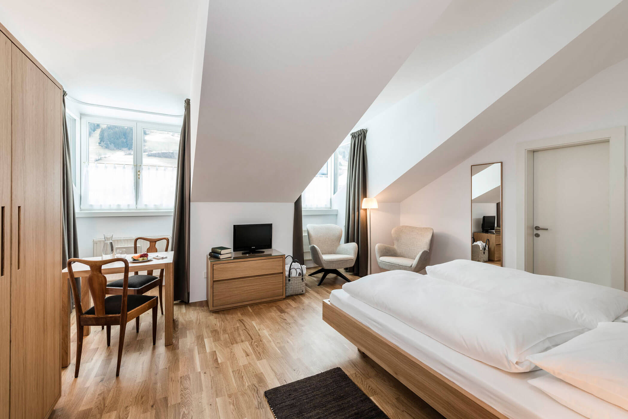 Bright attic bedroom with a double bed, table, chairs, TV, armchairs, and large windows.