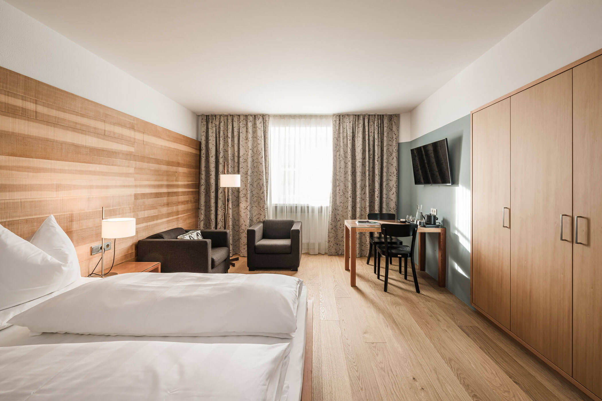 Modern hotel room with a double bed, armchairs, desk, TV, and large window with patterned curtains.