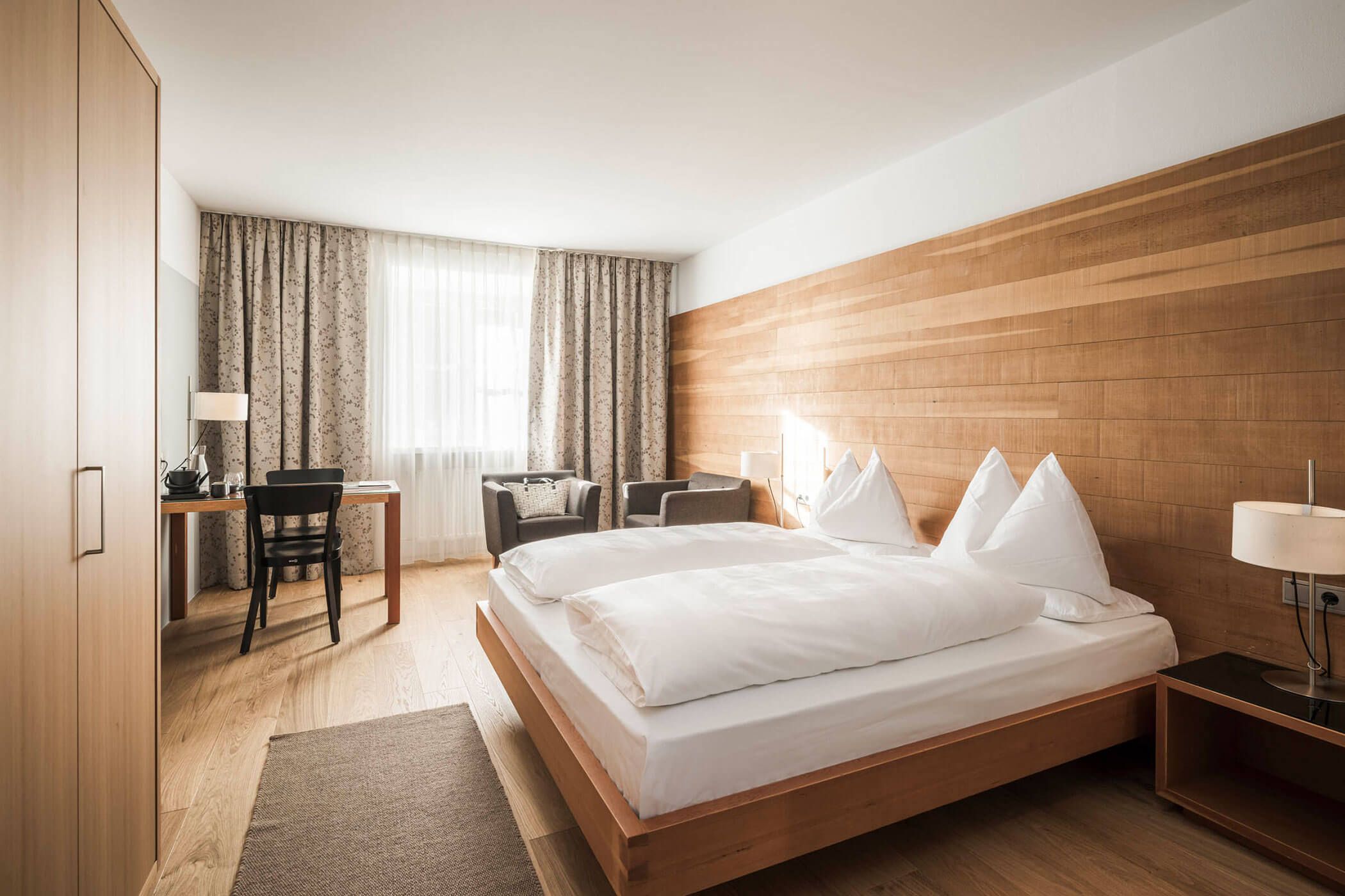 A modern hotel room with a double bed, desk, chairs, large window, and wooden accents.
