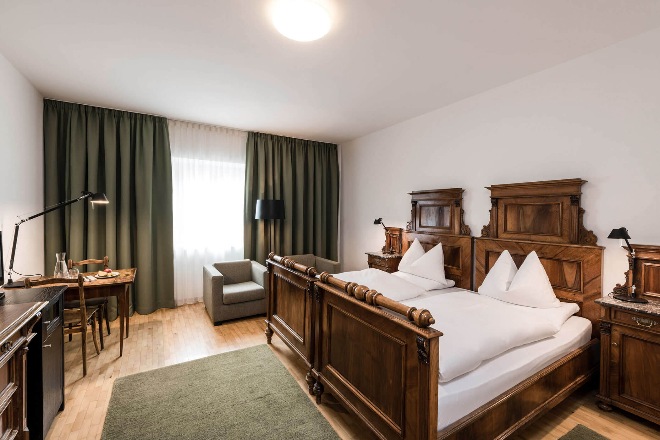 A cozy hotel room with antique wooden furniture, double bed, desk, armchair, and green curtains.