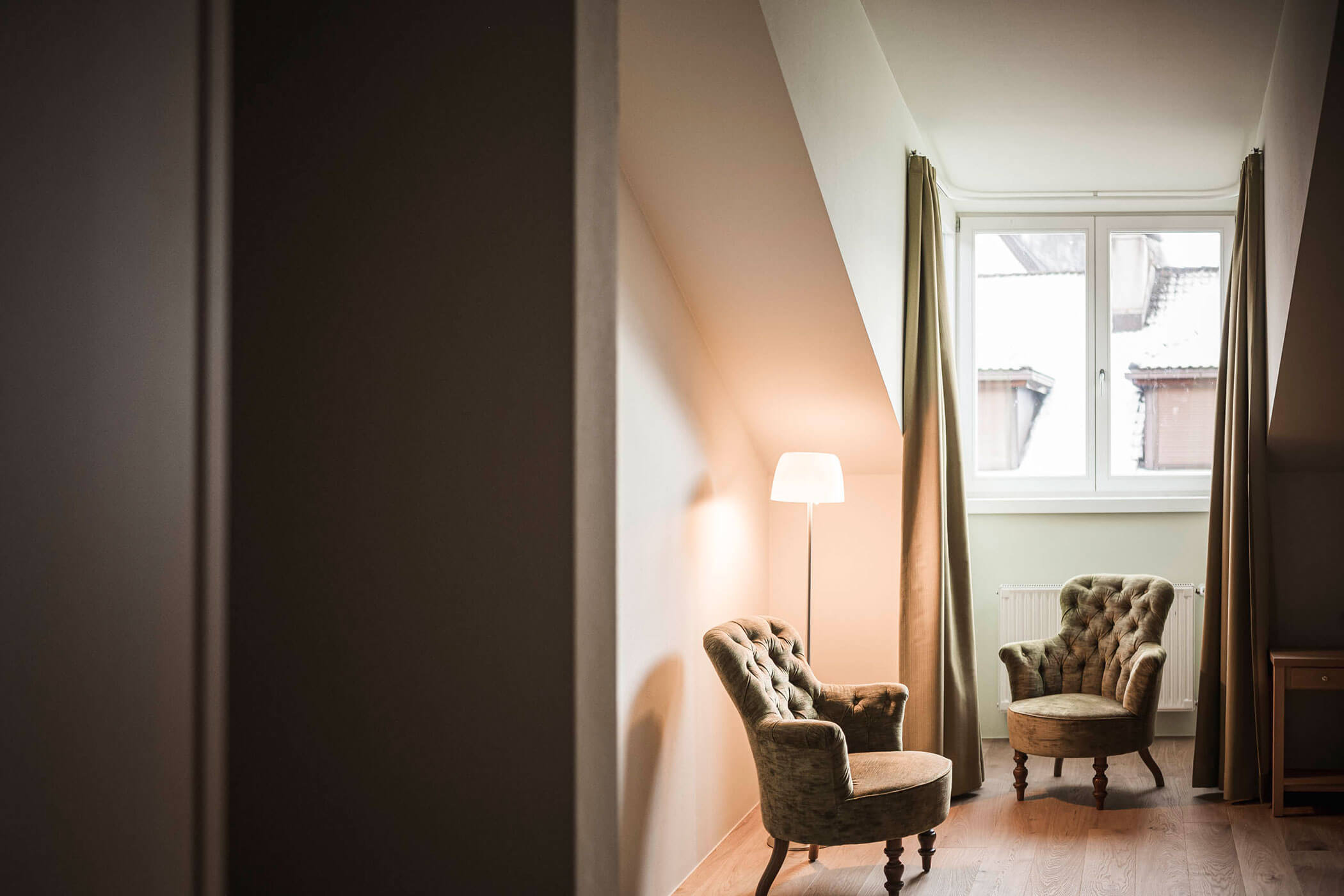 Two vintage armchairs and a floor lamp by a window in a cozy, softly lit nook with slanted ceilings.