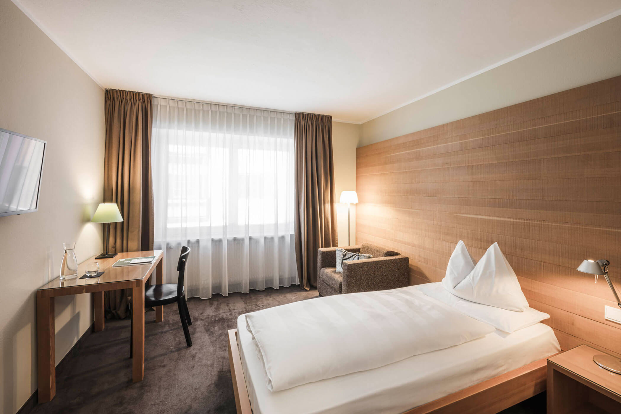 A modern hotel room with a single bed, desk, chair, armchair, lamp, and large window with curtains.