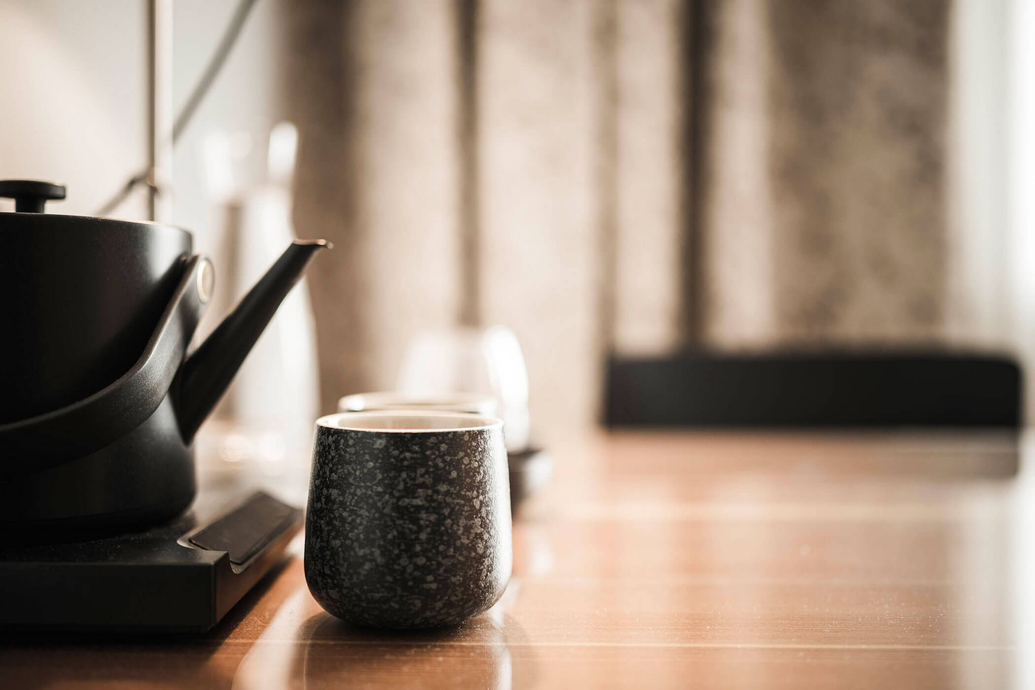 A black teapot and a speckled black cup sit on a wooden table in soft, natural light.