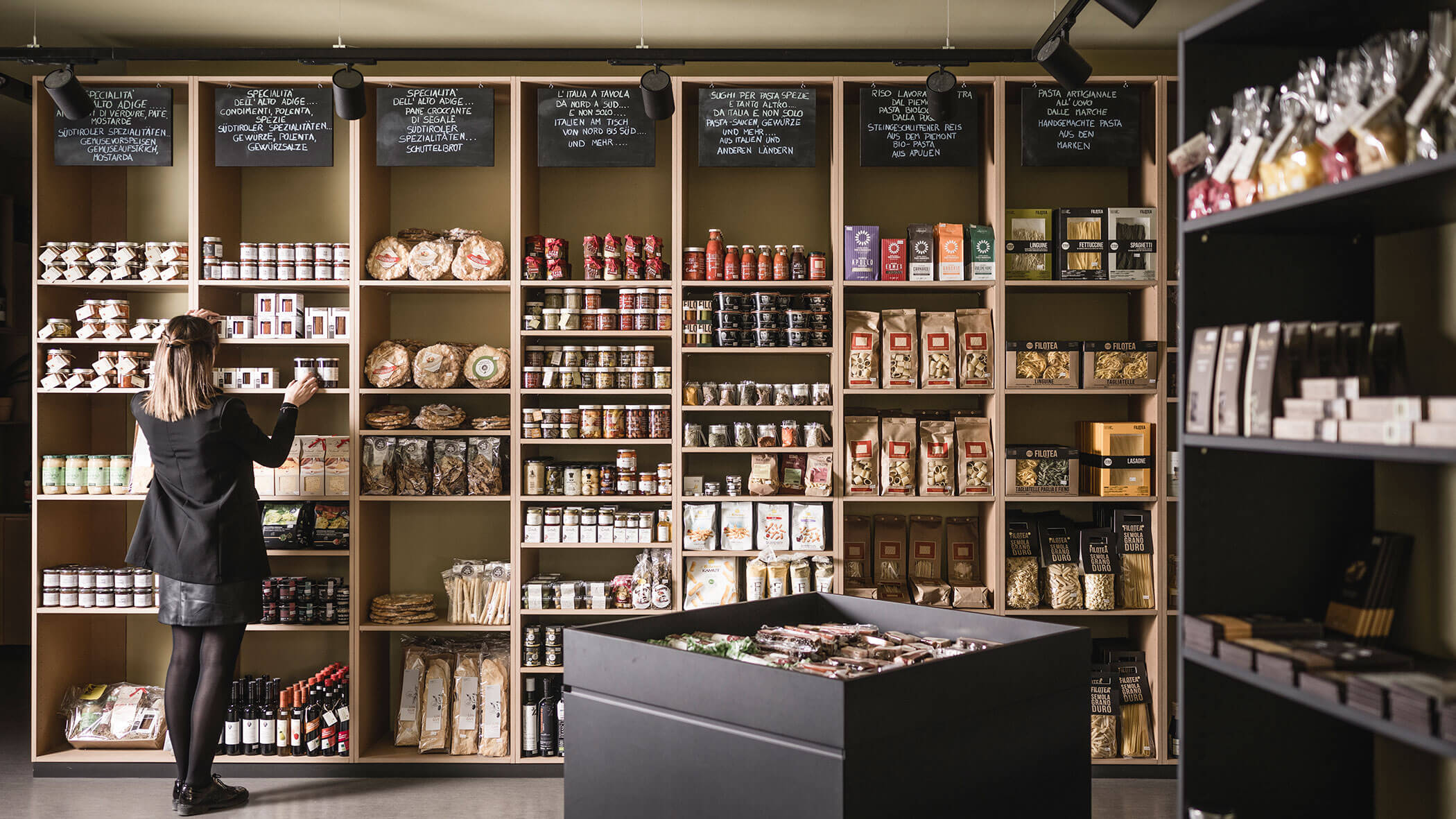 A woman shops in a gourmet grocery store with shelves of packaged food and chalkboard menus above.