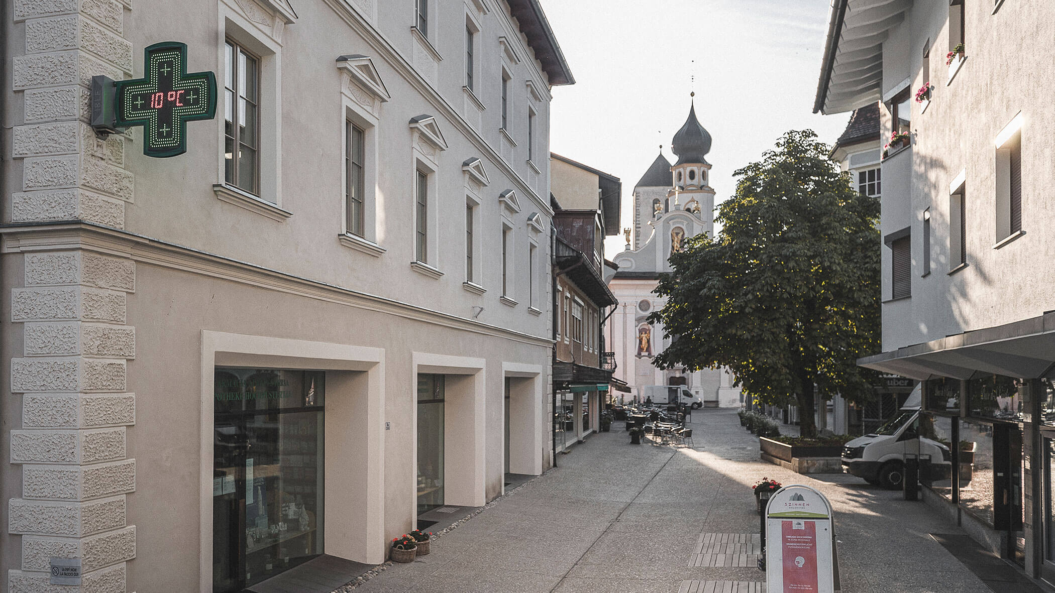A quiet European street with a pharmacy sign, shops, and a church with domed towers in the background.