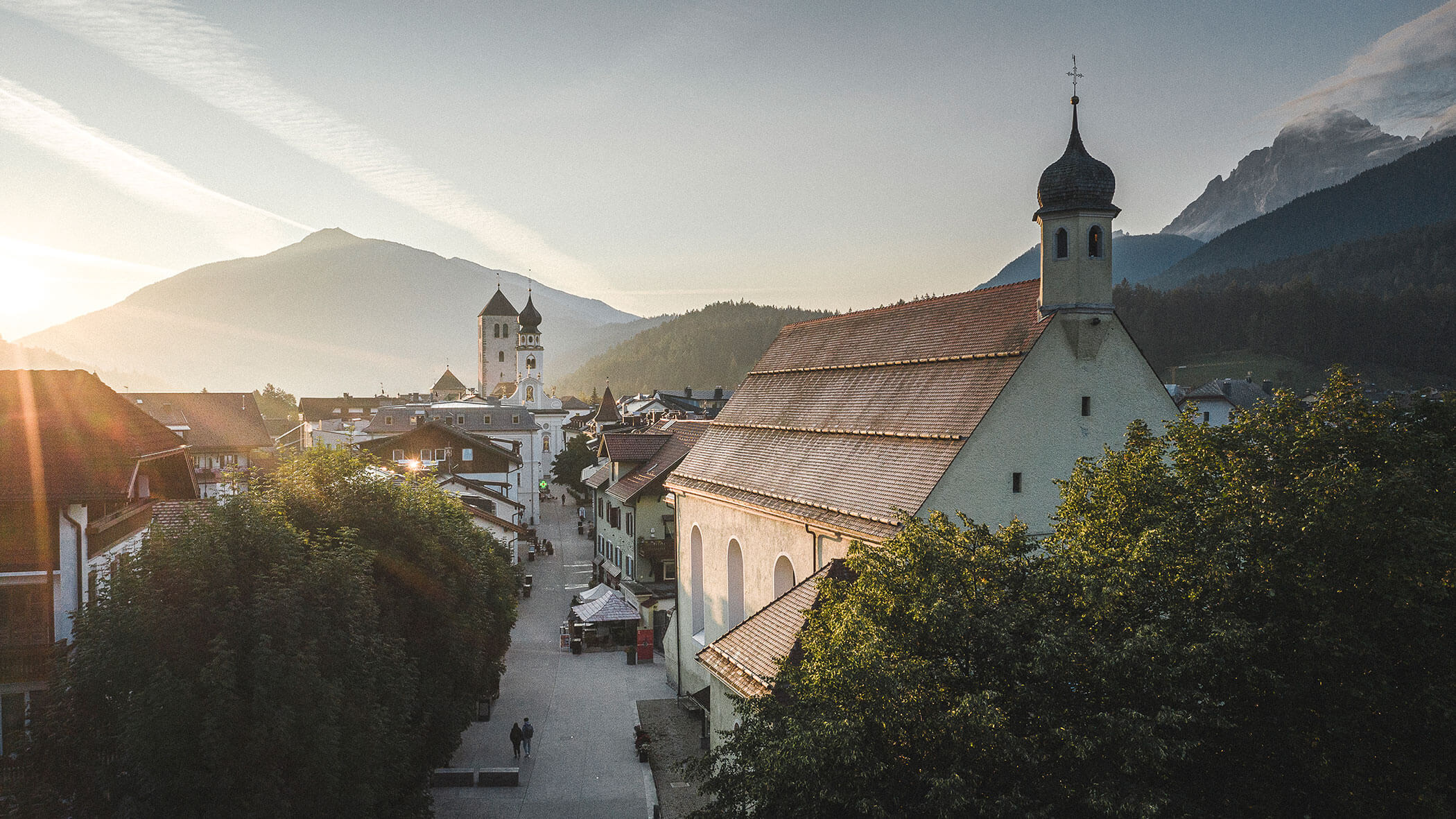 A quaint village with churches, mountains, and trees at sunrise, with sunlight streaming across the scene.