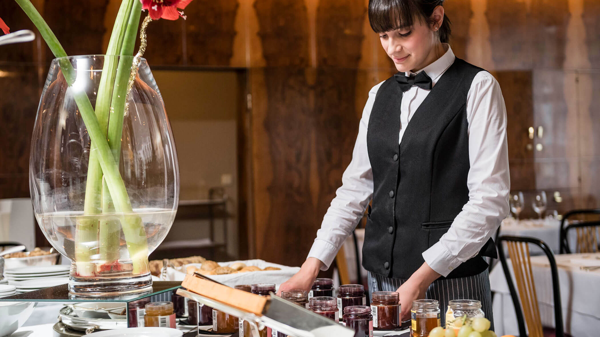 A server in uniform arranges glass jars of jam on a table in a restaurant dining area.