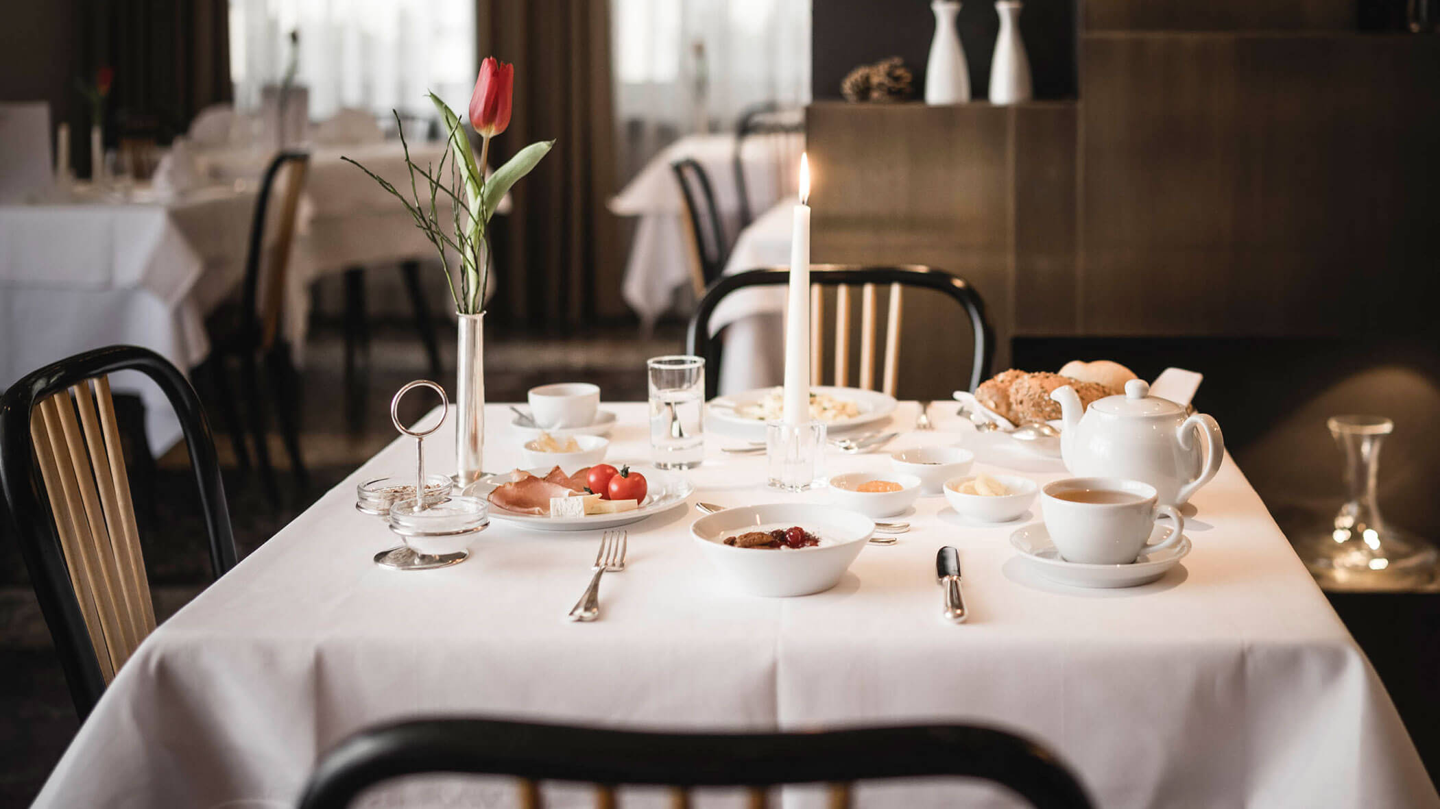 Elegant breakfast table with white dishes, a lit candle, a red tulip, and assorted food items.
