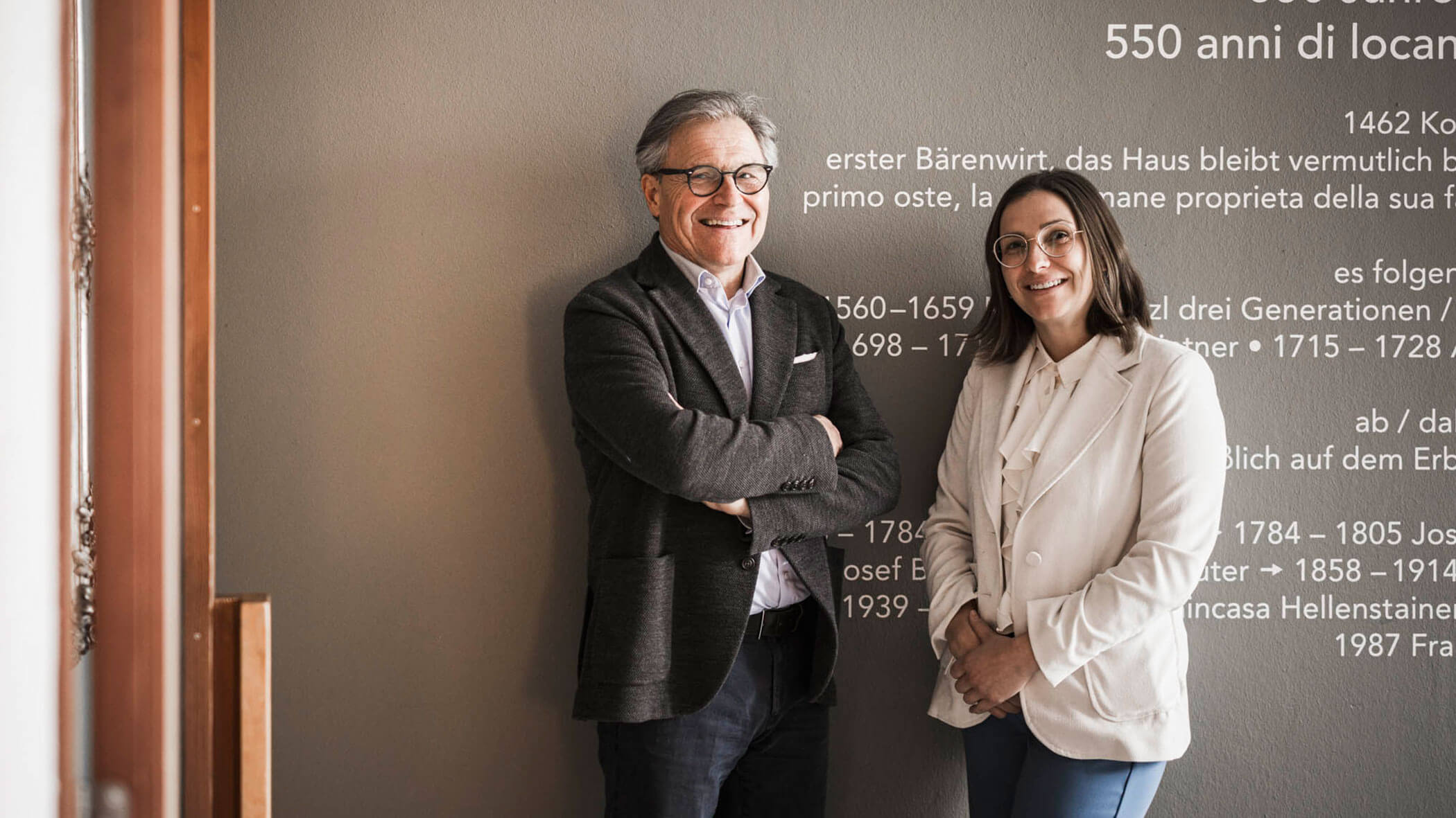 Two people in business attire smile while standing against a grey wall with historical text written in Italian and German.