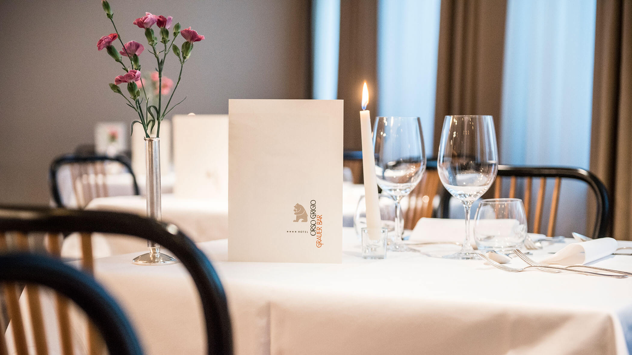 Elegant table setting with a candle, menu, wine glasses, and pink flowers in a vase on a white tablecloth.