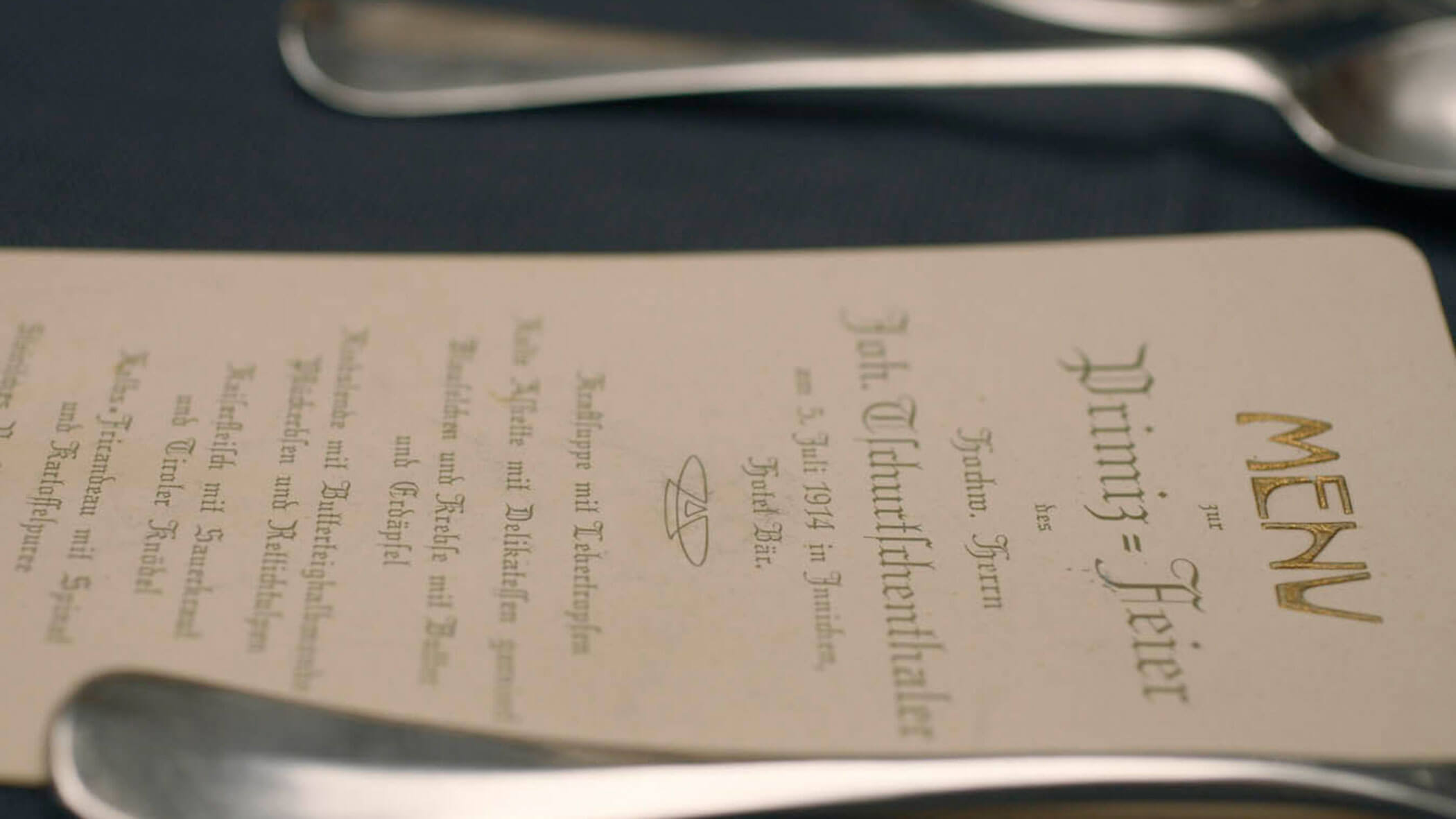 A close-up of a German menu with silver cutlery on a dark tablecloth.