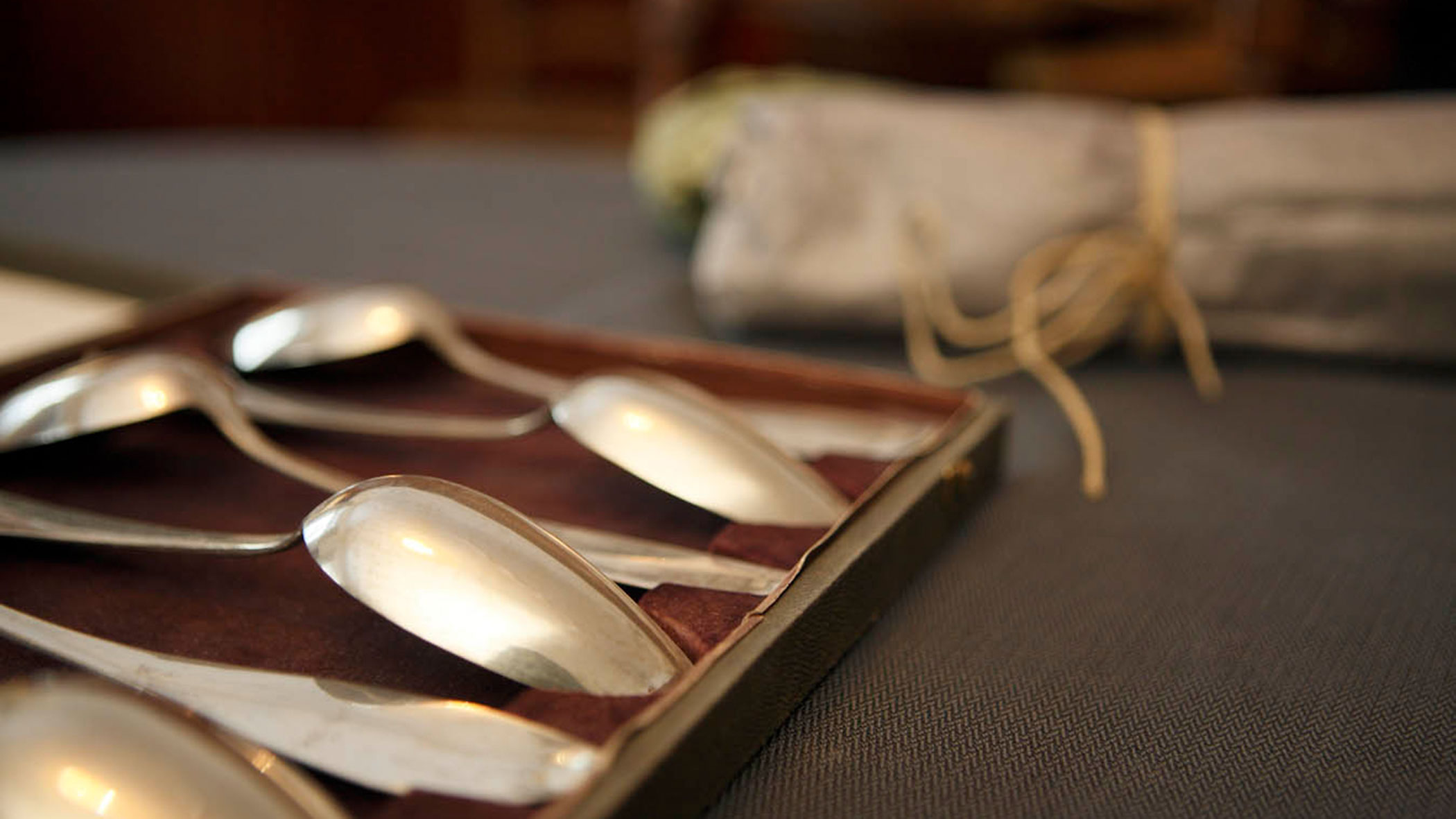 Close-up of silver spoons in a box on a table, with a rolled napkin tied with string in the background.