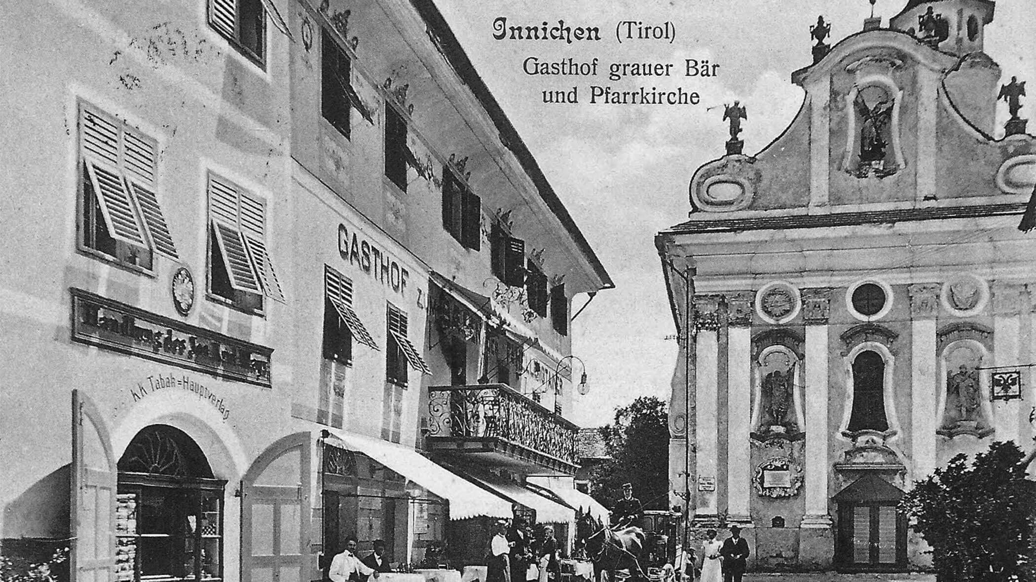 Black and white photo of an inn and a church in Innichen, Tirol, with people gathered outside.