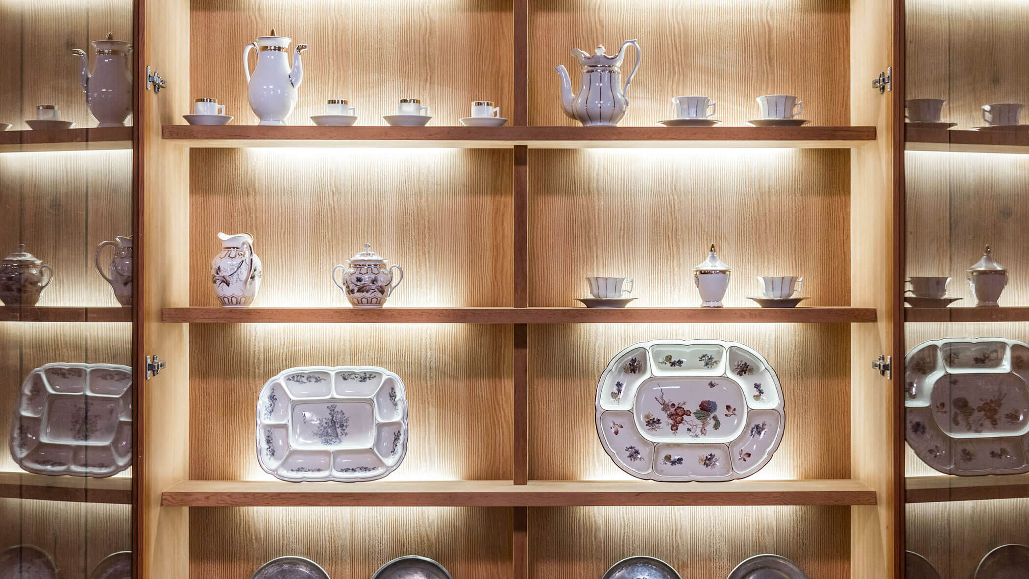 Lit wooden shelves displaying decorative china teapots, cups, and ornate plates in a showcase.